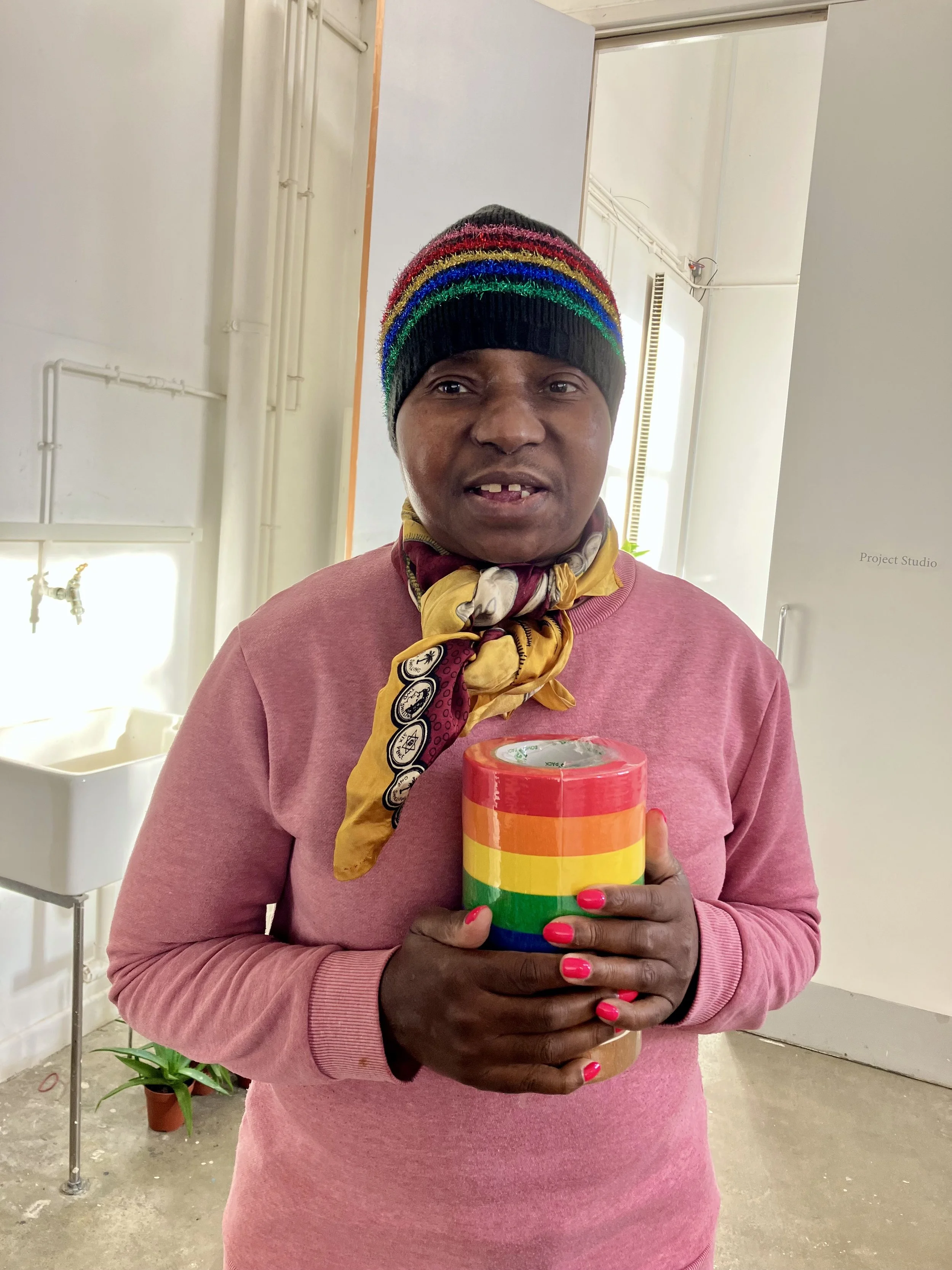 Portrait of a woman wearing a rainbow-striped hat and scarf, holding a roll of multicoloured tape, with a studio sink and door visible in the background