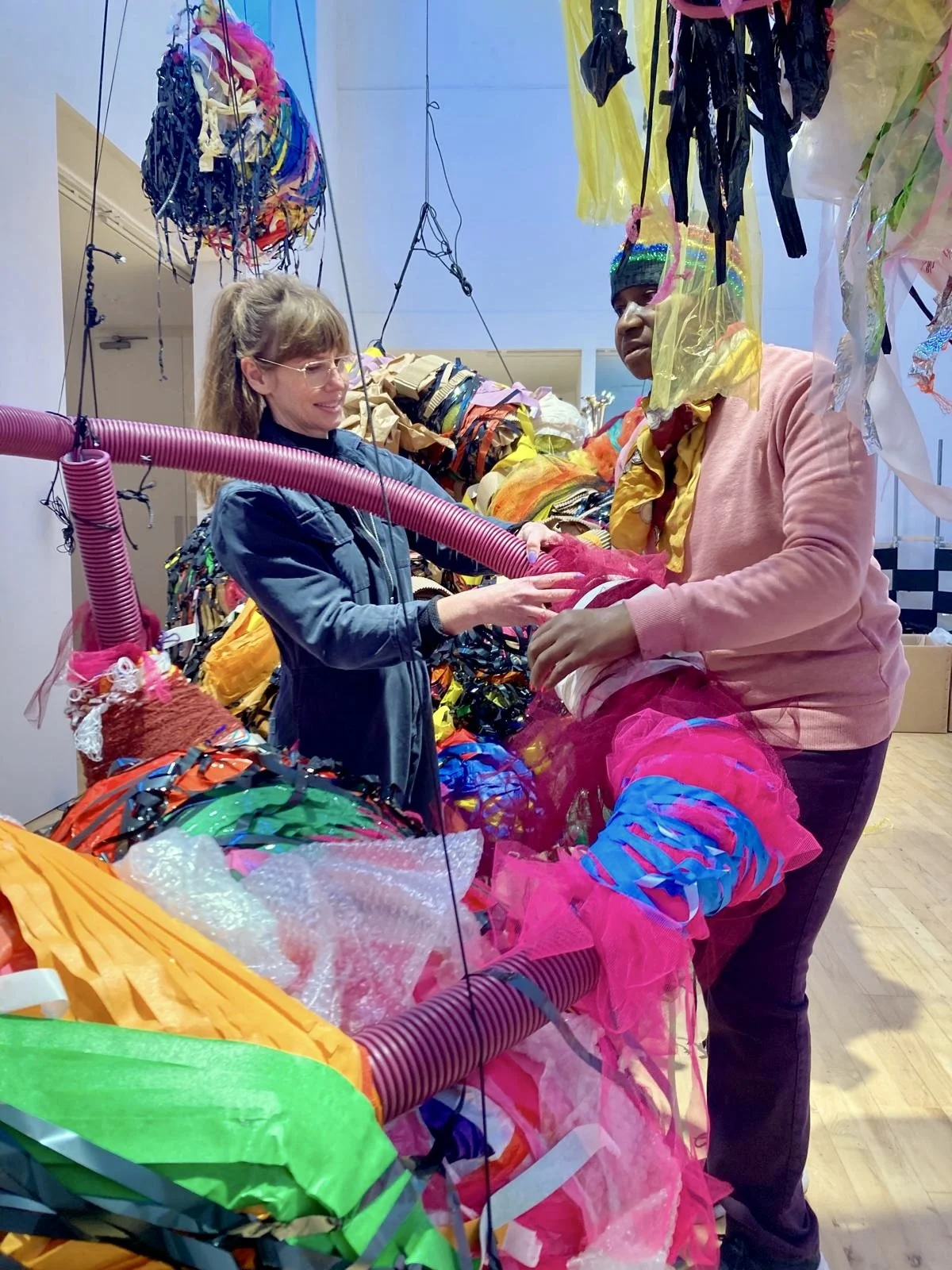 Two people stand within the installation, working together to wrap and secure bright pink and multicoloured materials around a suspended sculptural structure.