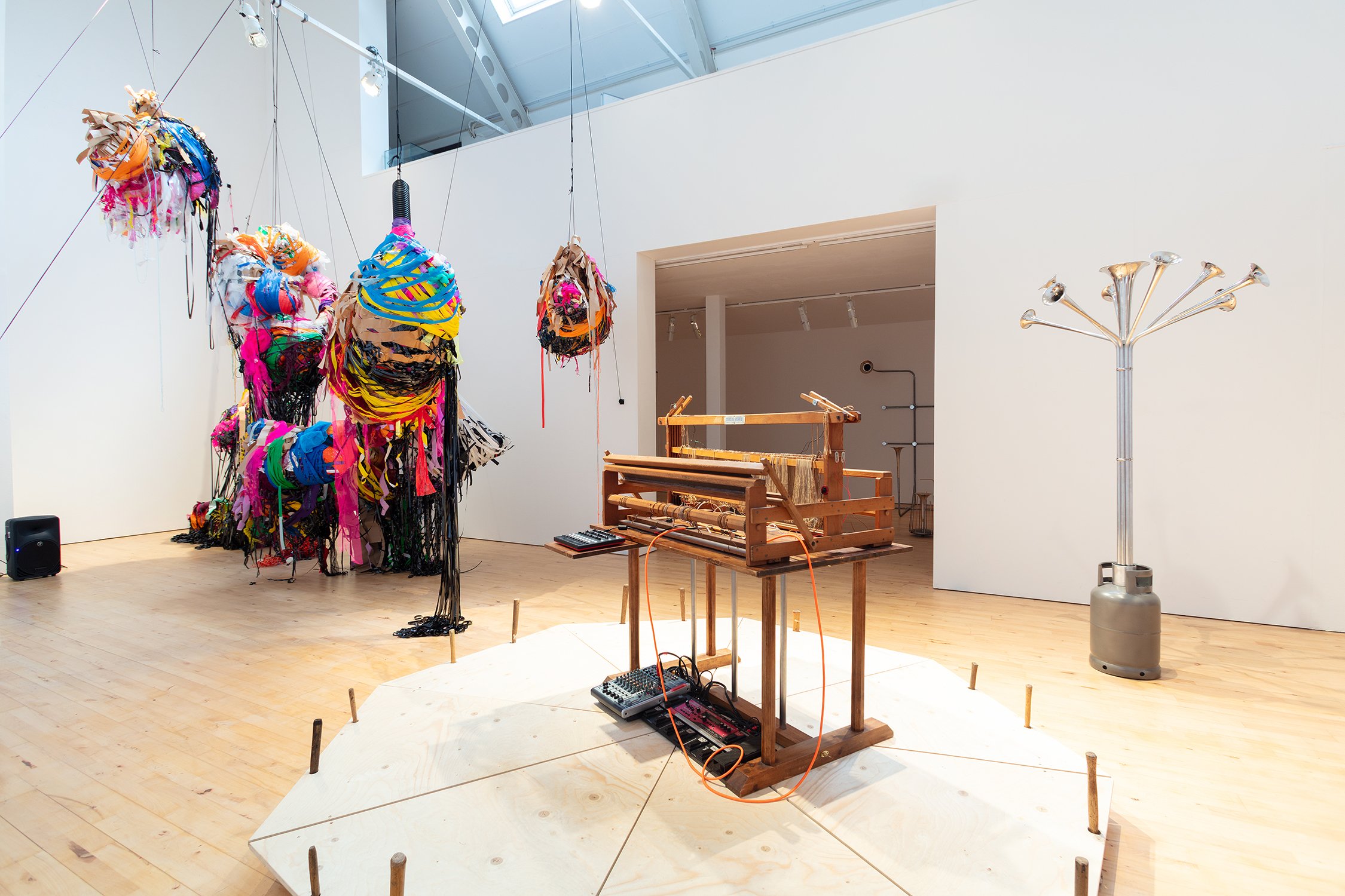 Wide gallery view of suspended, brightly coloured sculptural forms made from layered ribbons, plastics, and textiles, hanging from the ceiling; a wooden loom with audio equipment sits in the foreground.
