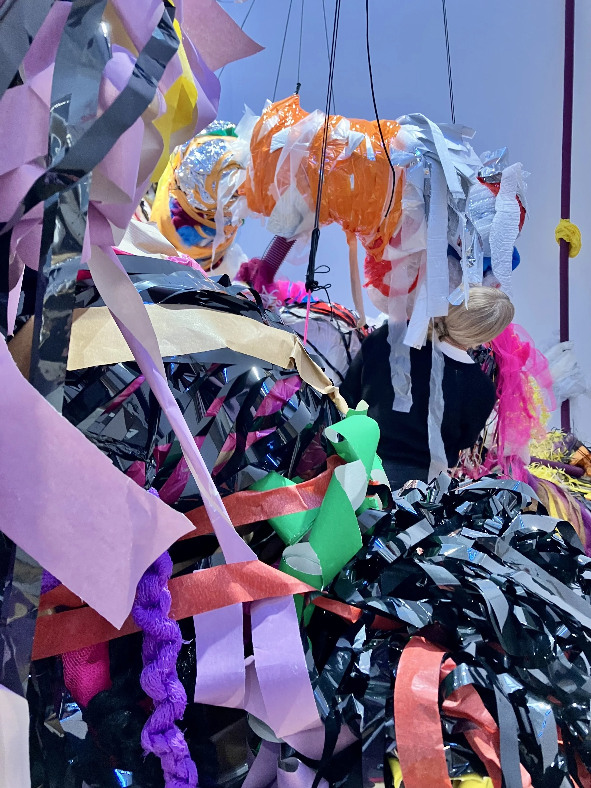 View from within the installation, showing dense layers of coloured plastic and fabric strips in the foreground, with a person working among the suspended forms in the background.