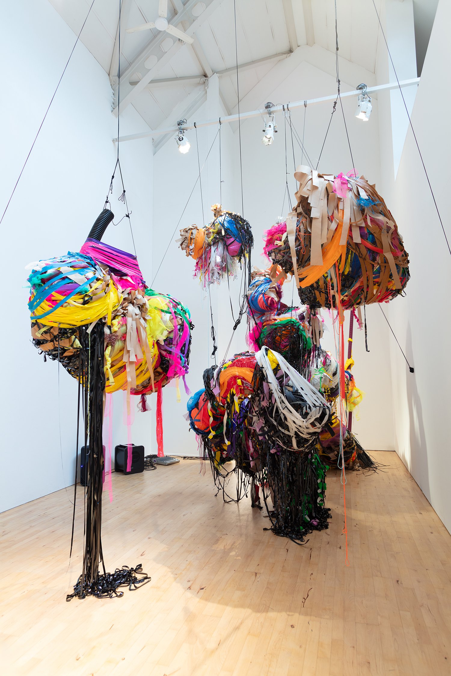 Closer view of suspended sculptural forms, highlighting dense layering of colourful materials and the contrast between bright ribbons and black trailing elements.