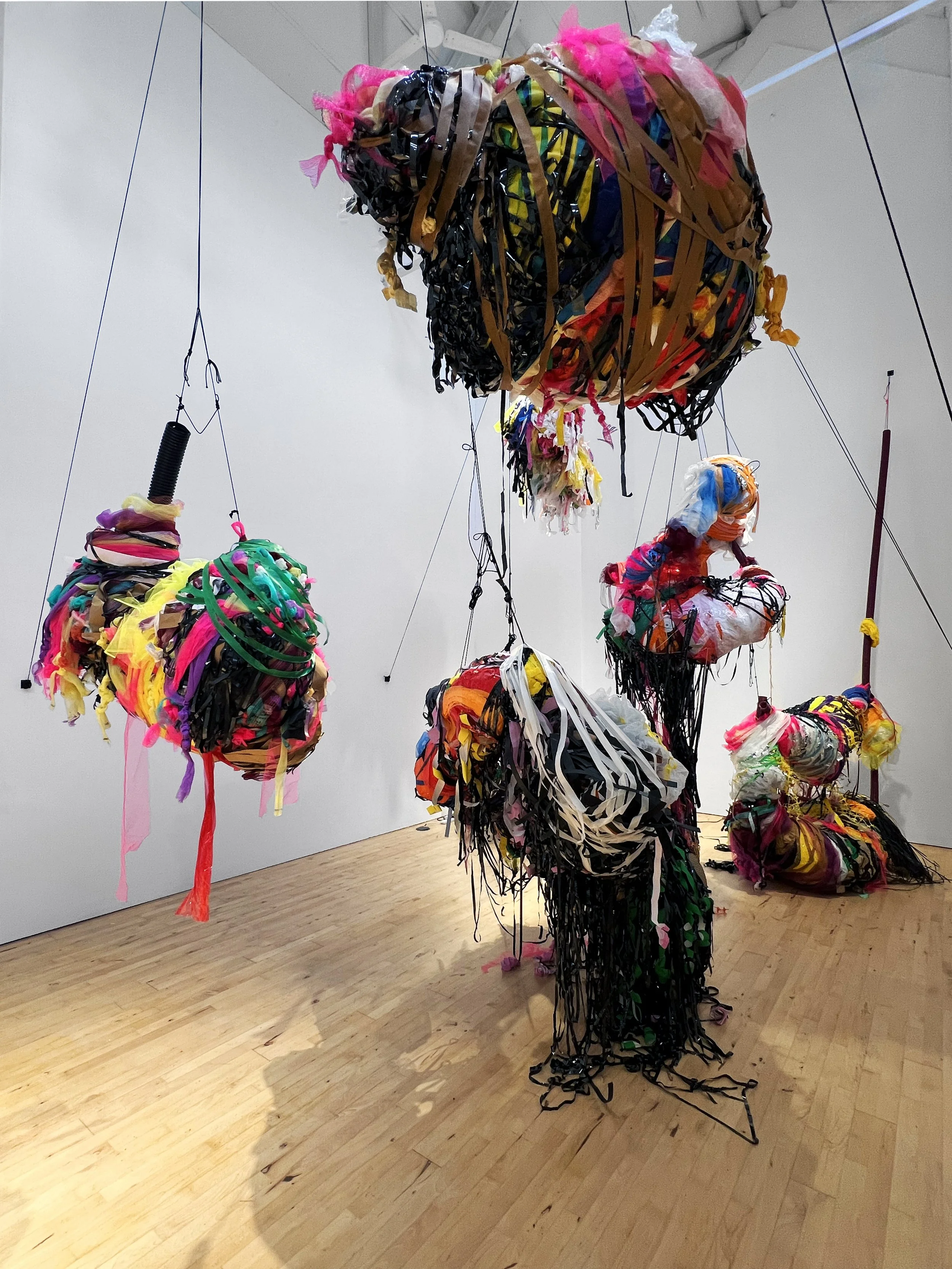 Installation view of suspended, multicoloured sculptural forms made from layered plastic, fabric, and tape, hanging at different heights in a white gallery space.