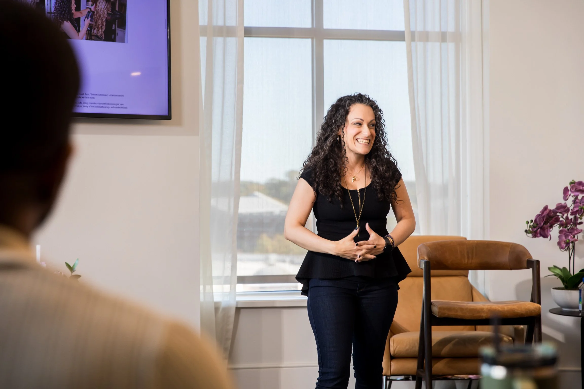 A woman with curly dark hair wearing a black top and pants, smiling, stands in a bright room with cream curtains and purple flowers on a table, giving a presentation or speech.