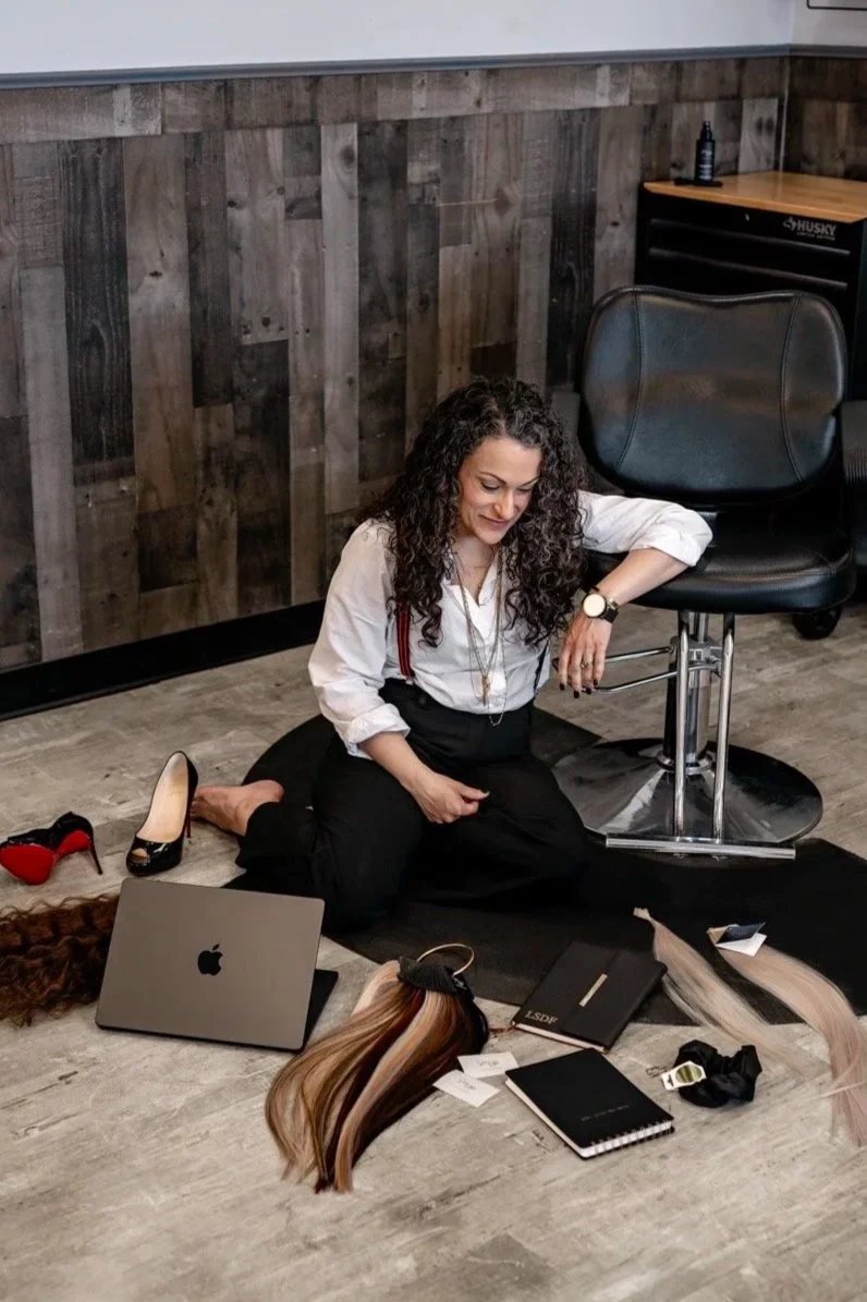 A woman with curly hair sitting on the floor in a salon, surrounded by hair swatches, a laptop, notebooks, and hair products.