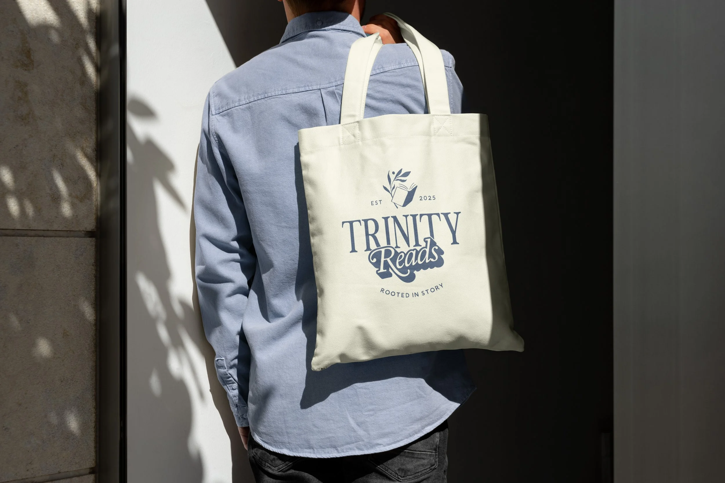 Person facing away carrying a white tote bag with 'Trinity Reads' logo and text, wearing a light blue shirt, standing next to a wall with shadowed foliage.