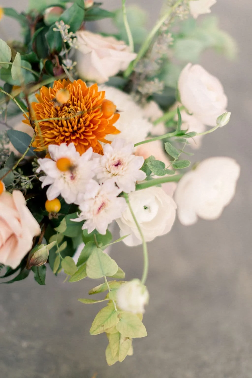 Designed by Twig & Bloom, this fine art floral arrangement offers an aerial view of delicate autumn blooms. A rich burnt-orange chrysanthemum brings a pop of fall color, beautifully paired with creamy white ranunculus, soft blush roses, and eucalyptu