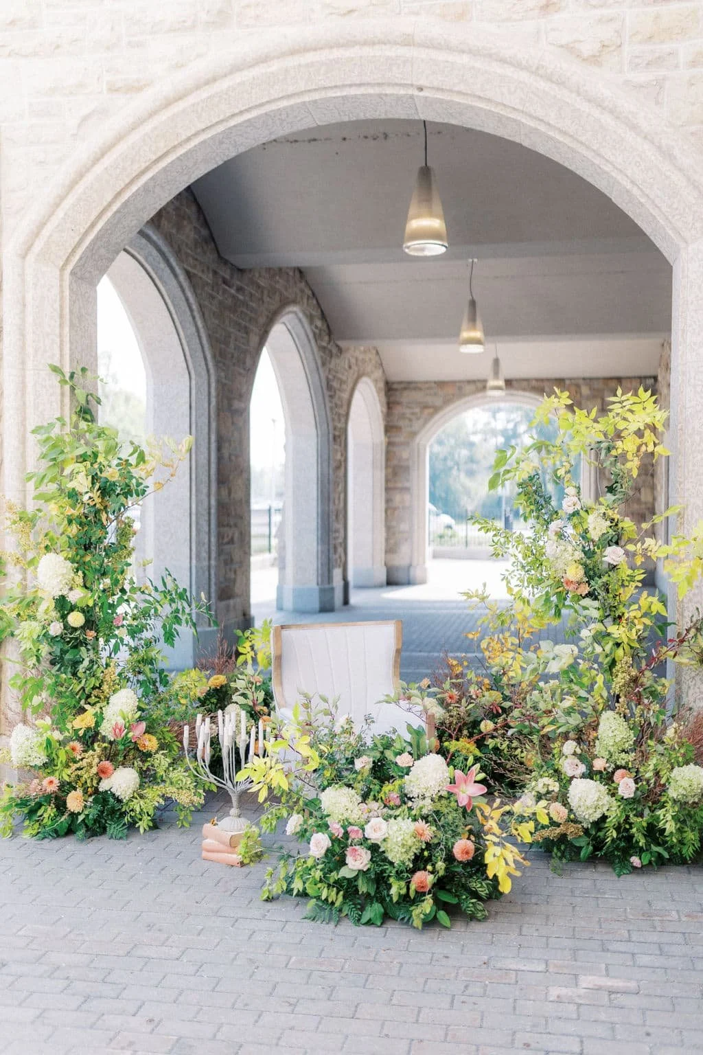 Framed by grand stone arches and elegant pendant lighting, this outdoor wedding ceremony setup features lush, ground-level floral installations and soaring floral pillars. Styled with a soft palette of yellow, blush, cream hydrangeas, and trailing gr