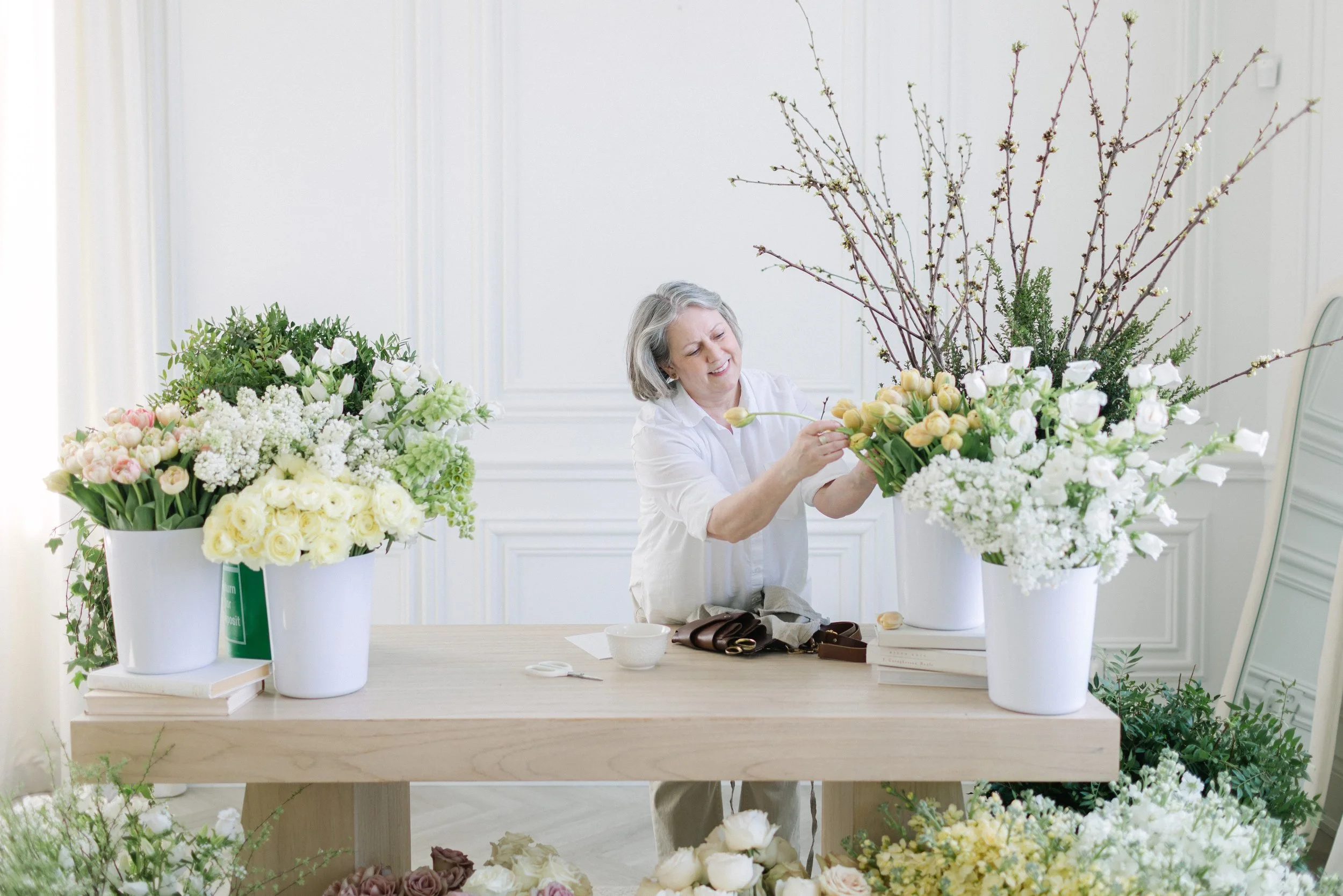 Twig & Bloom florist standing at a workbench, surrounded by Lilac, Tulips, Ranunculus, blossoming branches and other spring flowers in soft pastel colors.