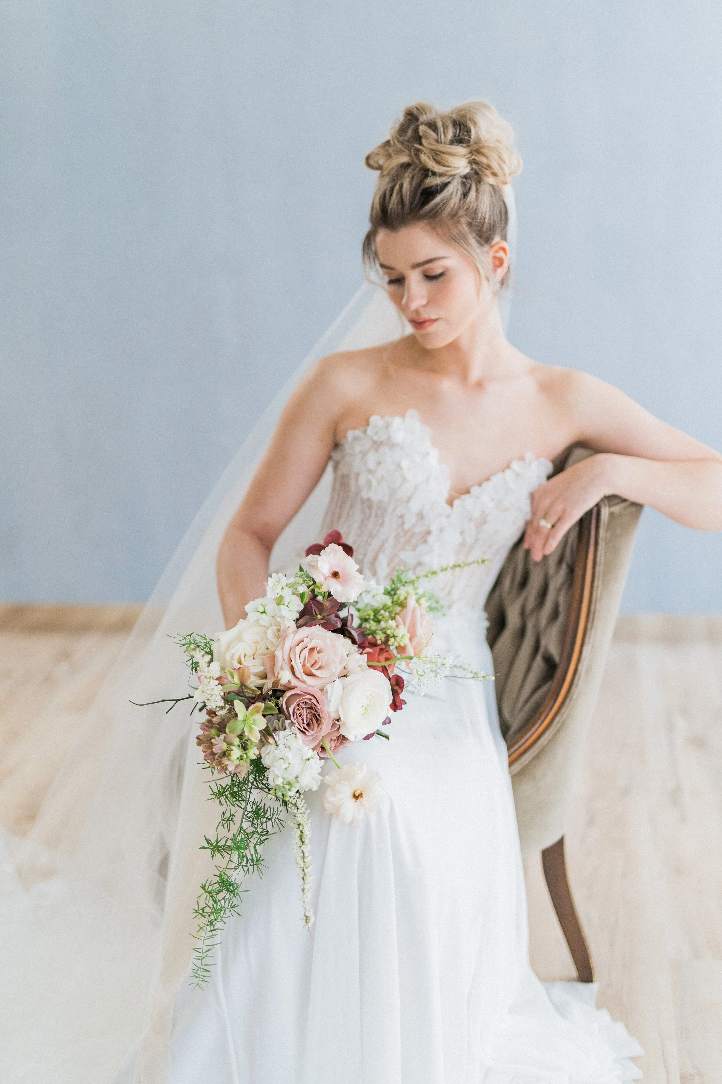 Bride with floral bouquet in elegant wedding dress seated on chair.