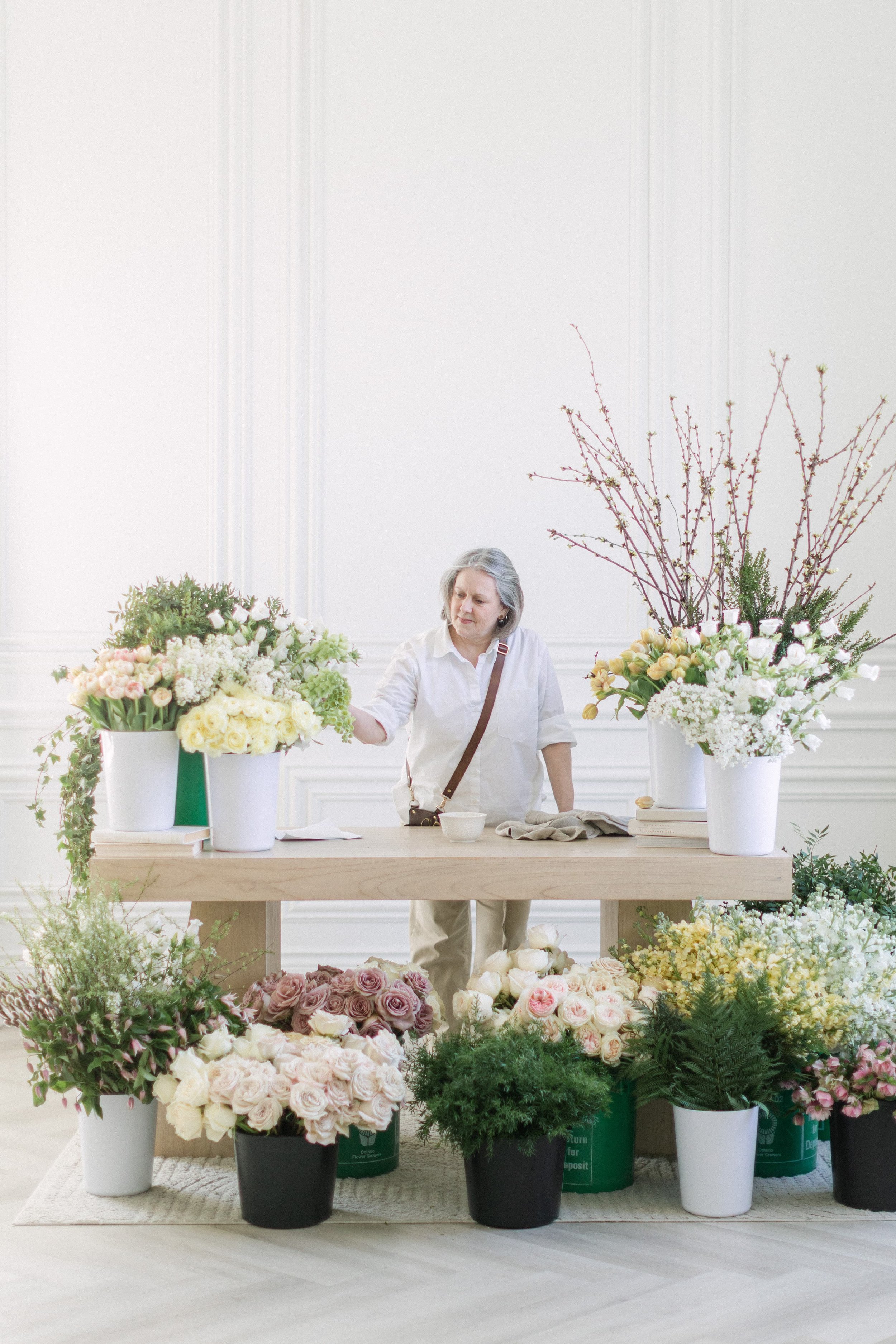 A florist from Twig & Bloom in Saskatoon is standing at a work table surrounded by spring flowers in pastel colors. She is preparing to create an arrangement.