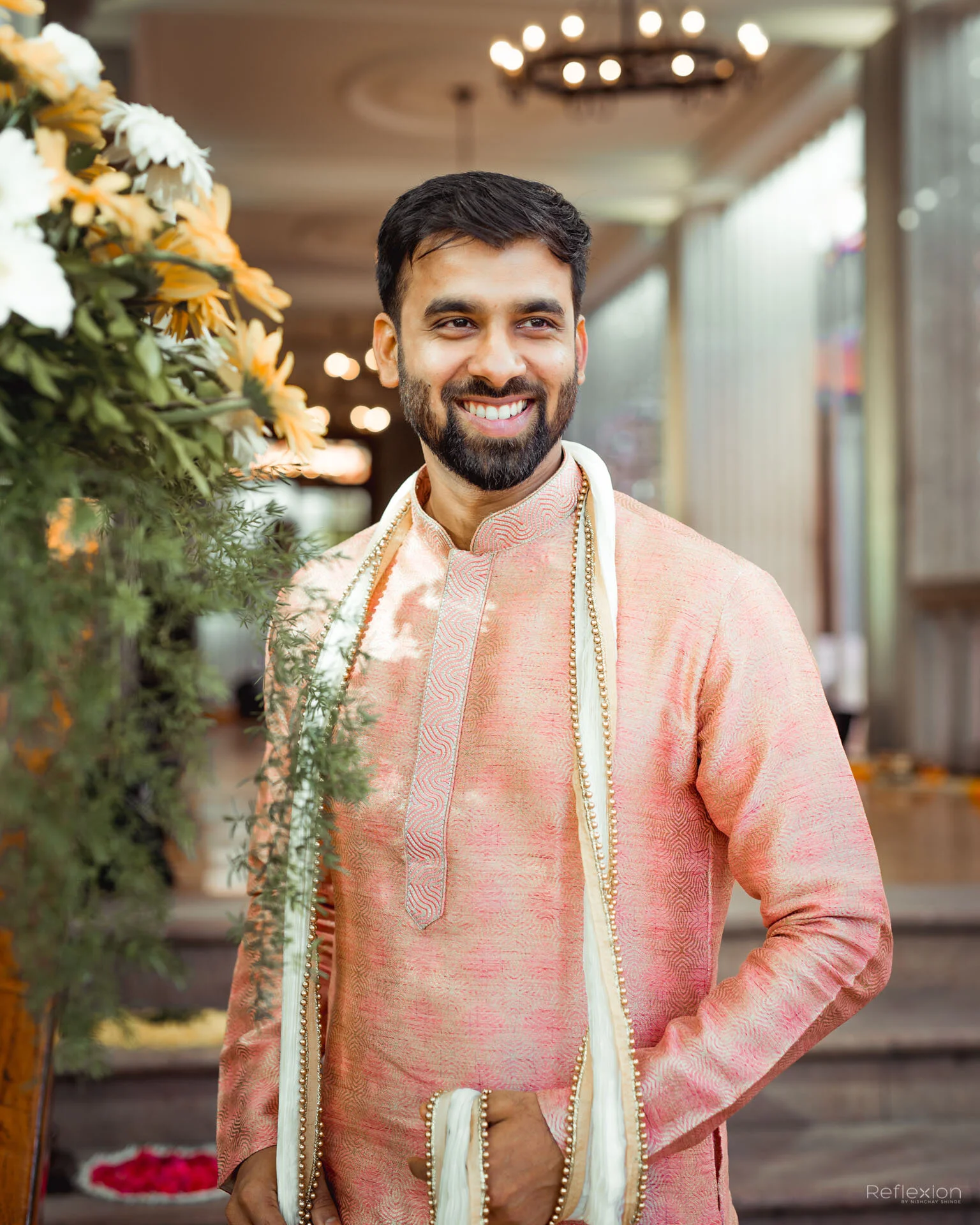 A man wearing traditional Indian attire, standing indoors, smiling, next to a floral arrangement.