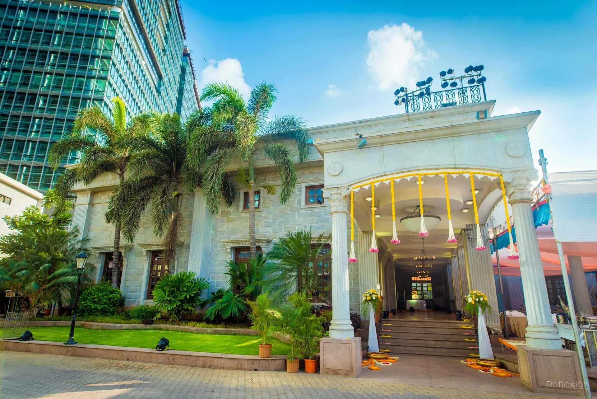 Luxurious hotel entrance decorated for celebration with flower garlands, potted plants, and steps leading to a grand door, surrounded by greenery and tall buildings under a blue sky.