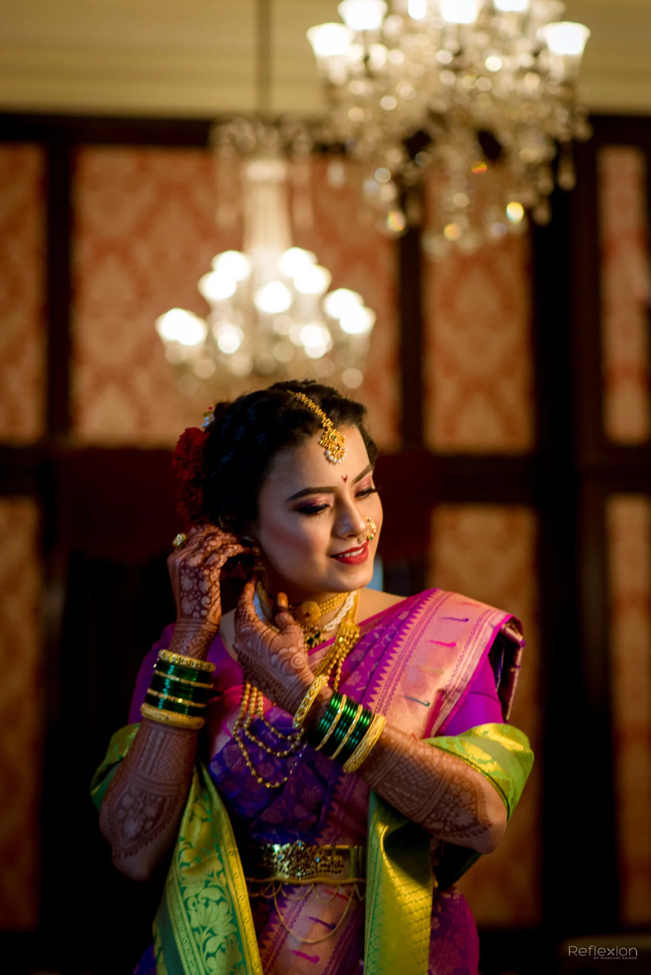 A woman dressed in traditional Indian attire, wearing vibrant colorful saree, numerous gold jewelry, and elaborate bangles, is fixing her earring in a decorated indoor setting with chandeliers in the background.