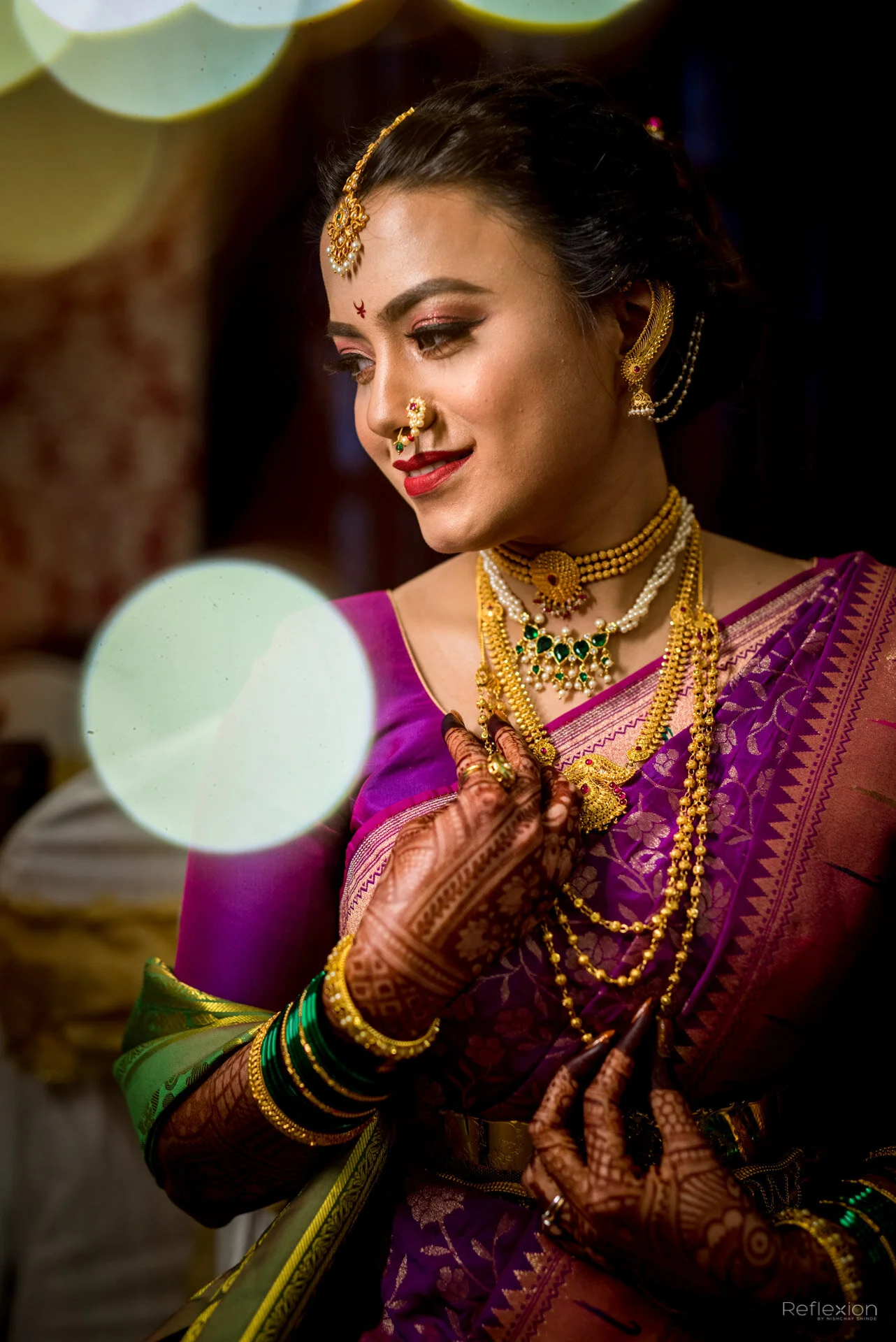A woman in traditional Indian attire with gold jewelry, a purple saree, and Mehndi on her hands, smiling and looking to her left.
