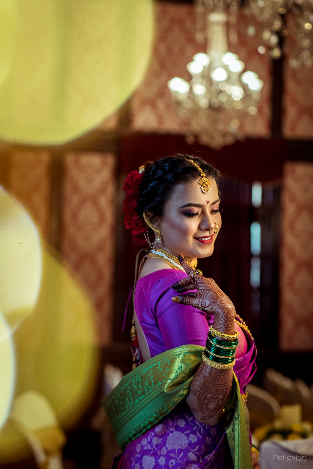 A woman dressed in traditional Indian attire, wearing purple and green saree, adorned with jewelry including bangles, earrings, necklace, and headpiece. She has henna on her hands and is smiling softly, with a blurred background of fabric and a chand