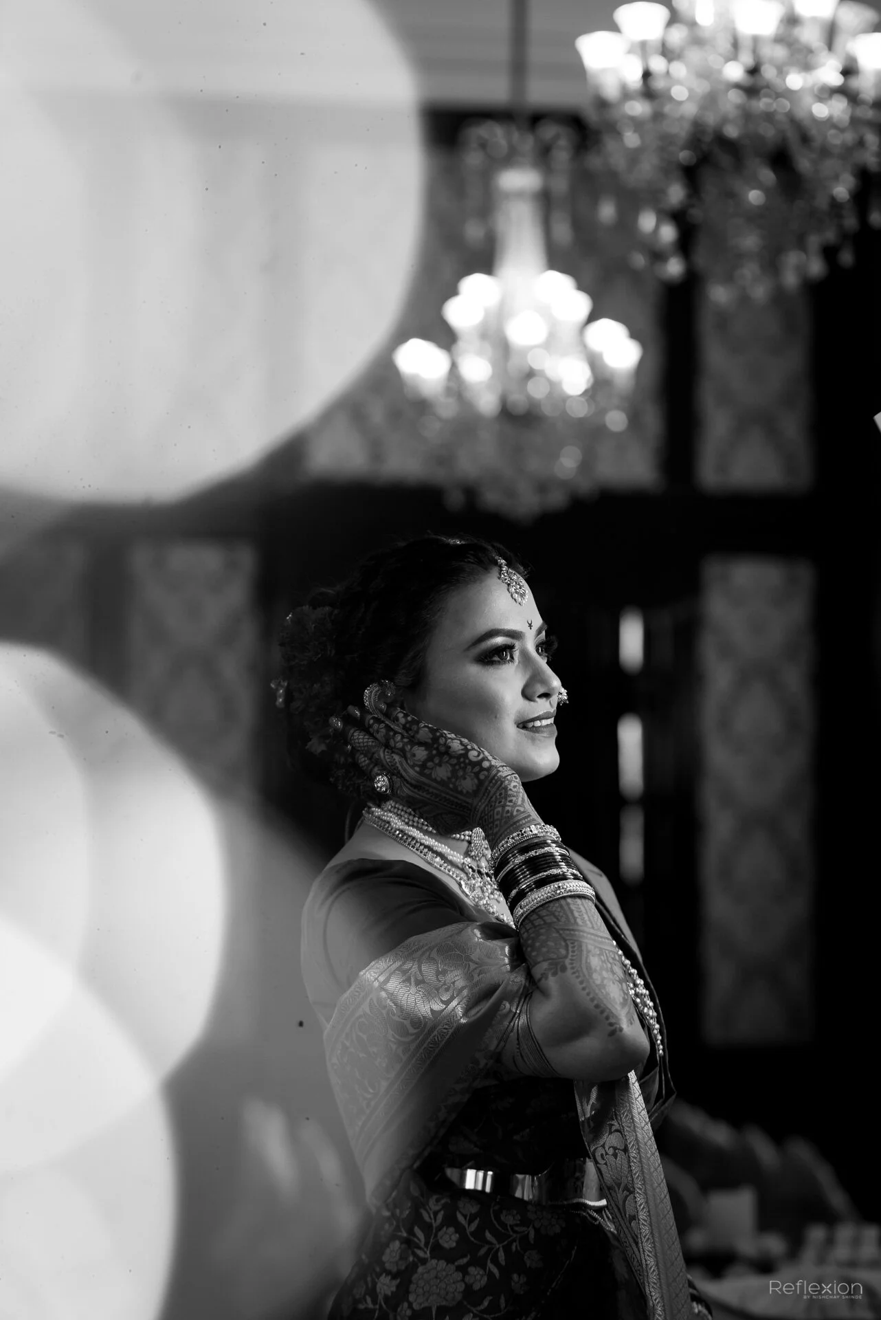 A woman dressed in traditional Indian attire, wearing jewelry and mehndi on her hands, smiling softly while adjusting her earrings.