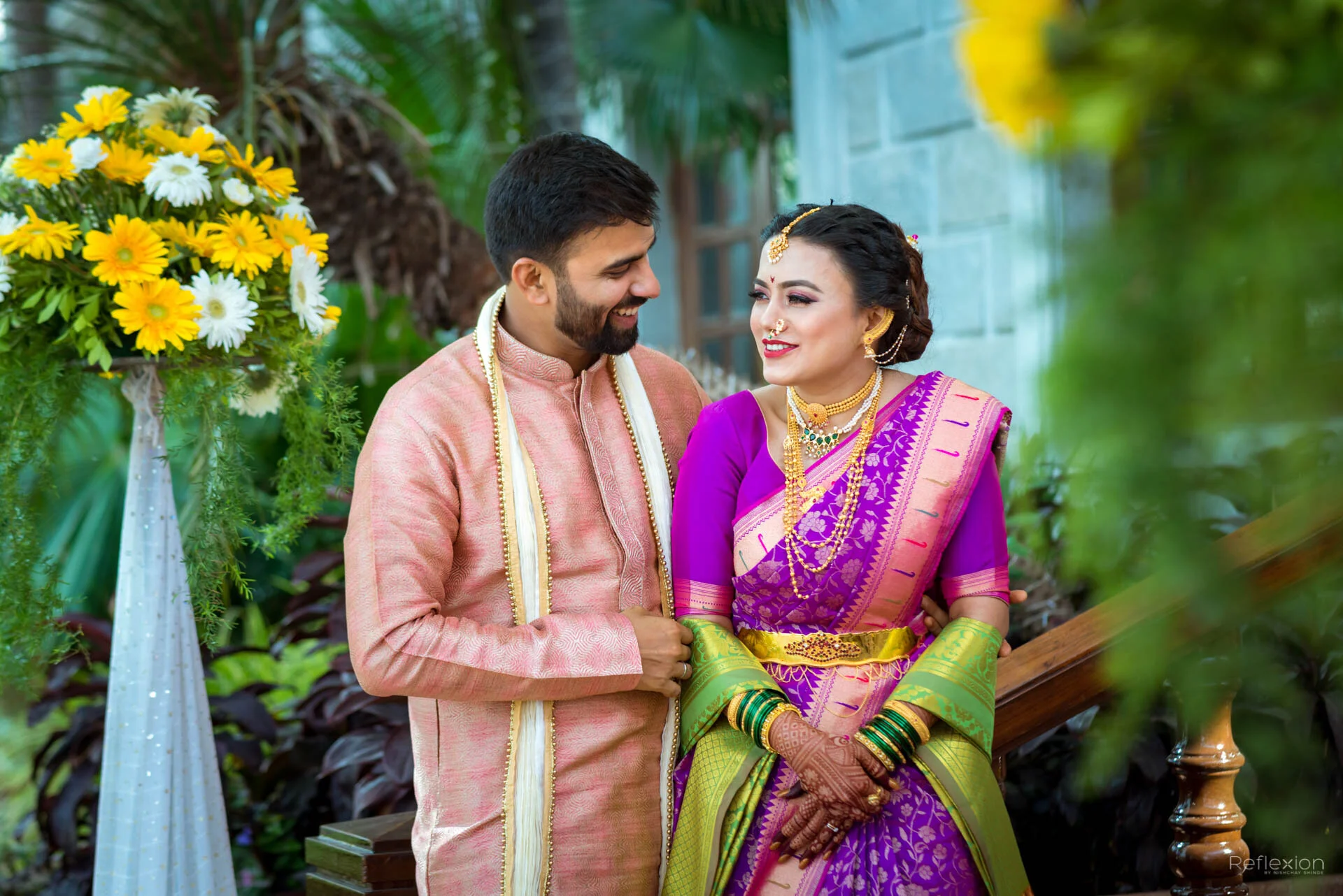 A couple in traditional Indian wedding attire smiling at each other outdoors, with flowers and greenery in the background.