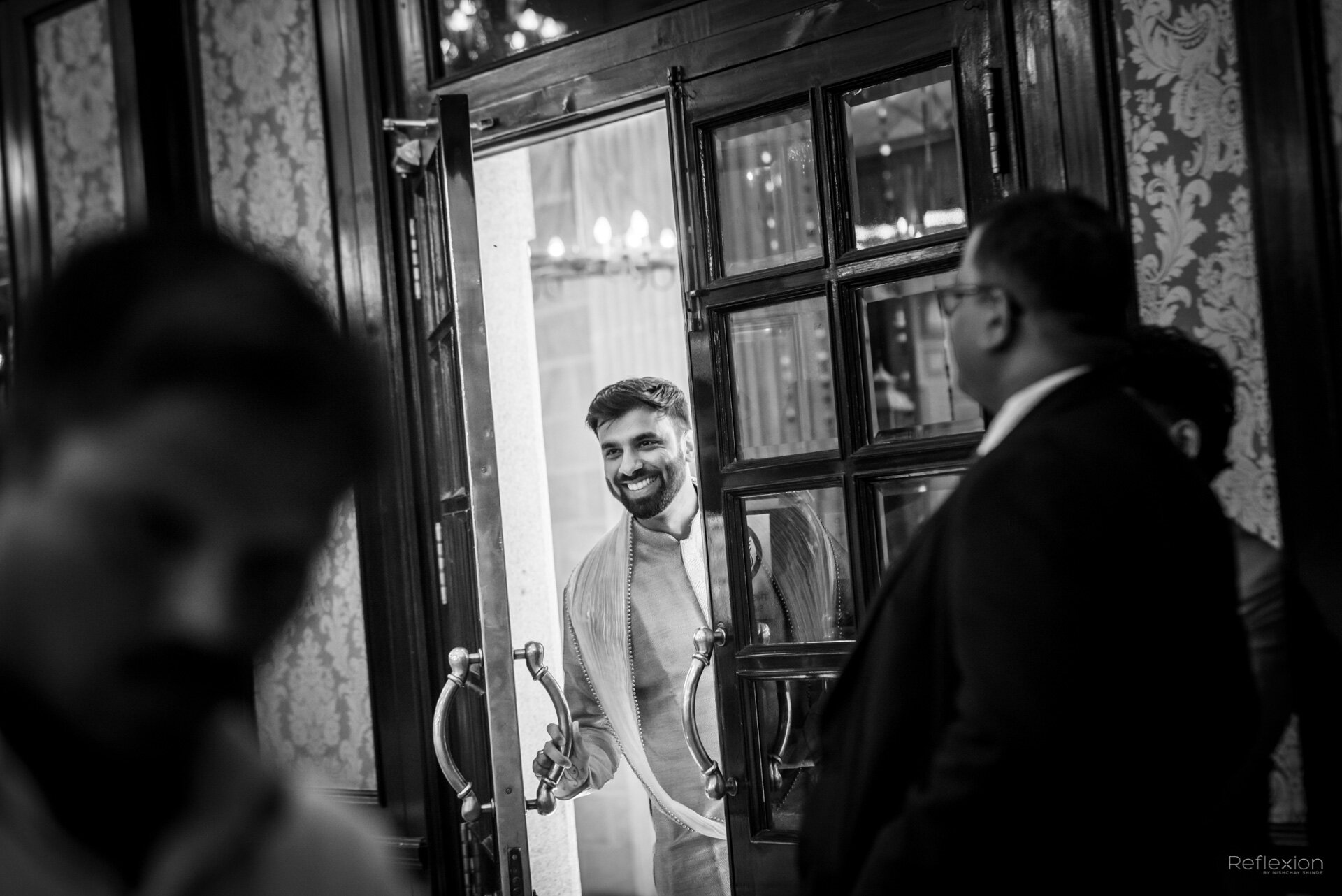 A man smiling as he opens a door for a group of people inside an elegant room with floral patterned wallpaper. The photo is in black and white.