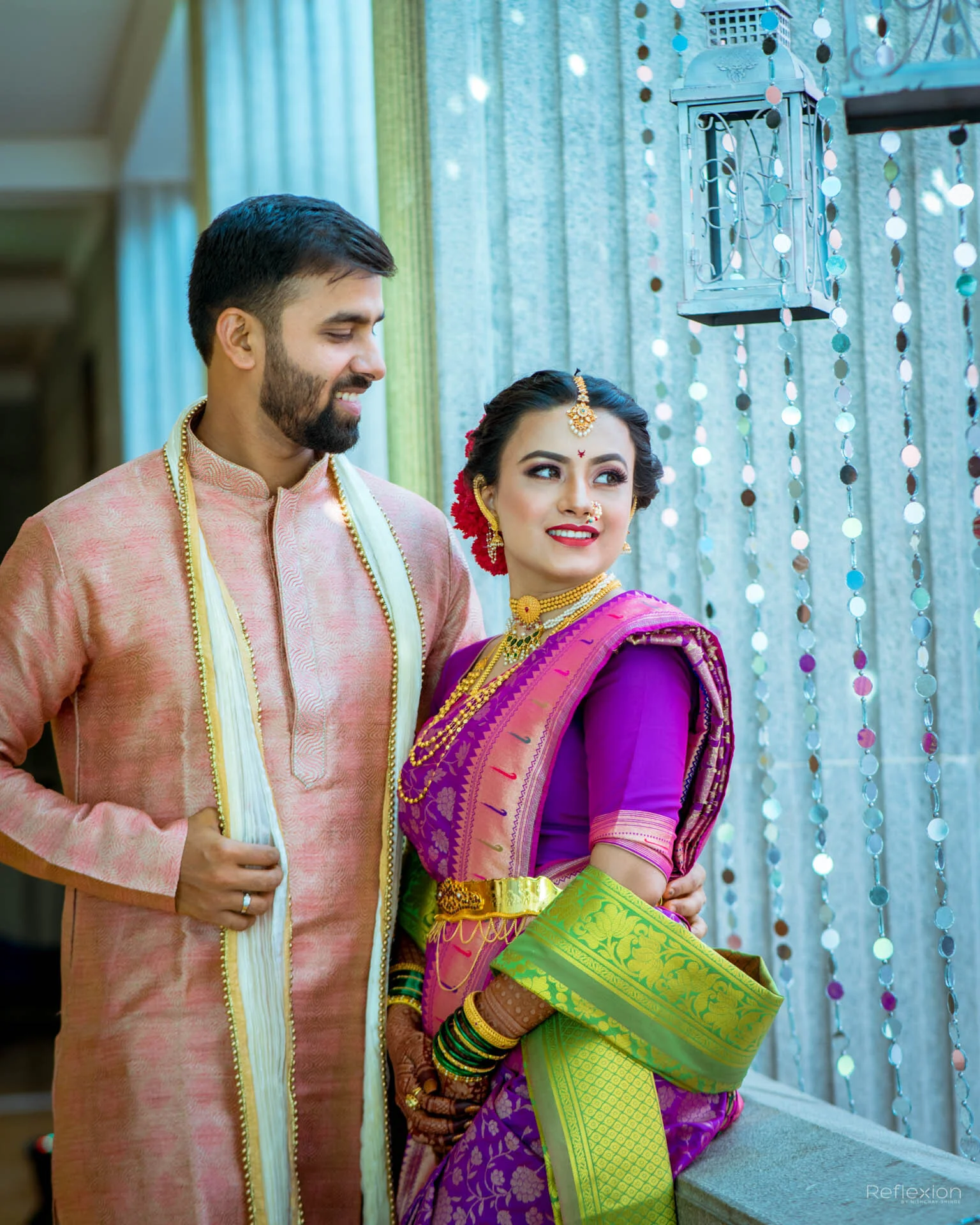 A couple dressed in traditional Indian attire, the woman in a purple saree with gold accents and jewelry, and the man in a pink kurta with a cream-colored dupatta, standing indoors near decorative curtains and hanging lanterns.