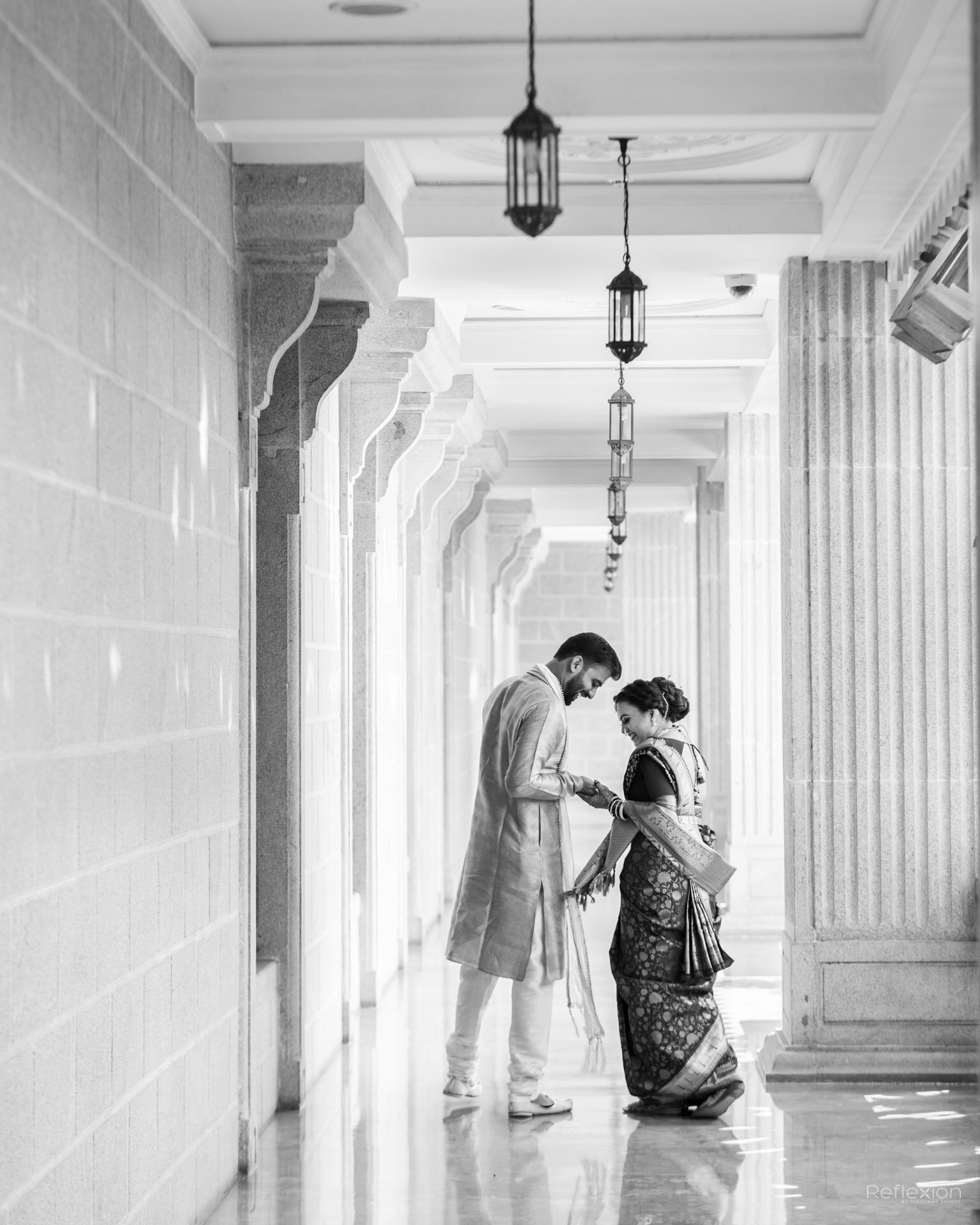 A man and woman dressed in traditional Indian wedding attire standing together in a corridor with high ceilings, hanging lanterns, and stone columns.