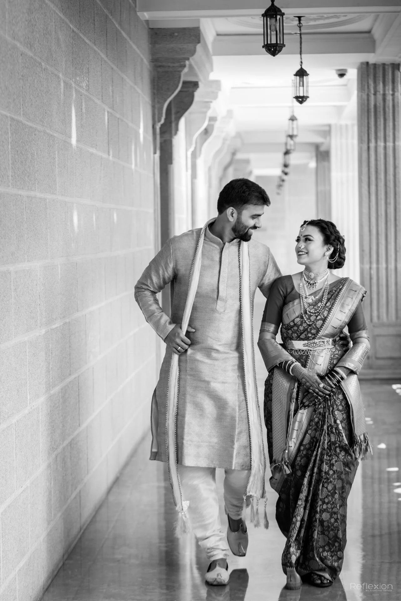 A man and woman dressed in traditional Indian attire walking together in a corridor, smiling at each other.