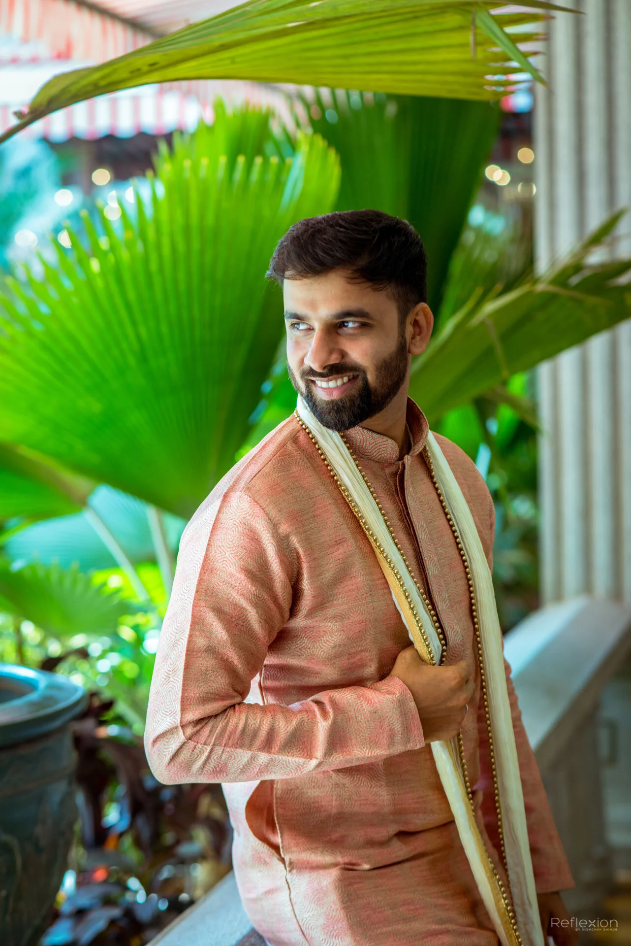 A man in traditional Indian attire standing outdoors among large green leaves, smiling and looking to the side.