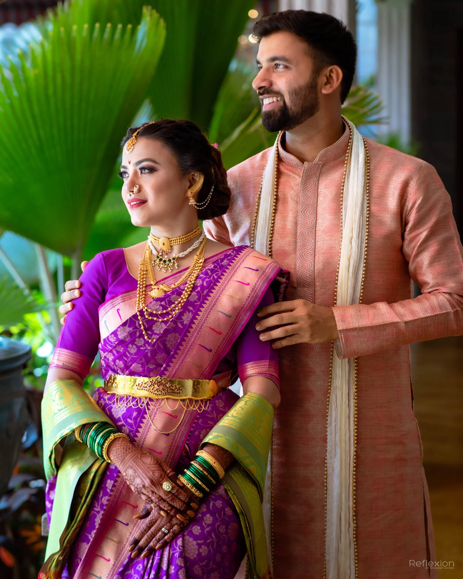 A couple dressed in traditional Indian wedding attire, standing outdoors with green palm leaves in the background. The woman is wearing a purple saree with gold jewelry, including necklaces, earrings, bangles, and a headpiece. The man is wearing a pe