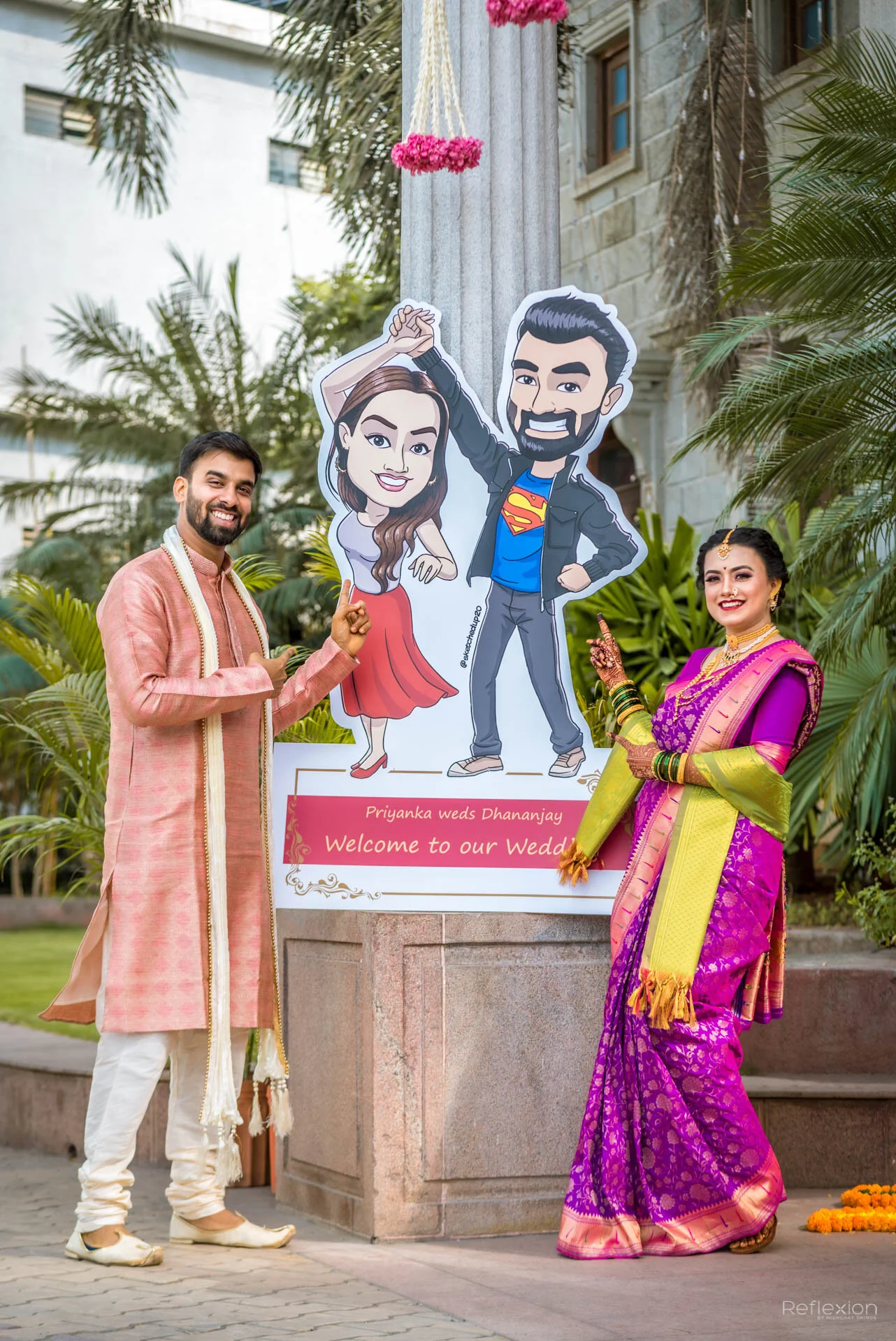 A man and woman in traditional Indian attire standing next to a wedding welcome sign with cartoon illustrations of themselves. The man points at the sign, and the woman smiles, both smiling at the camera amid lush greenery.