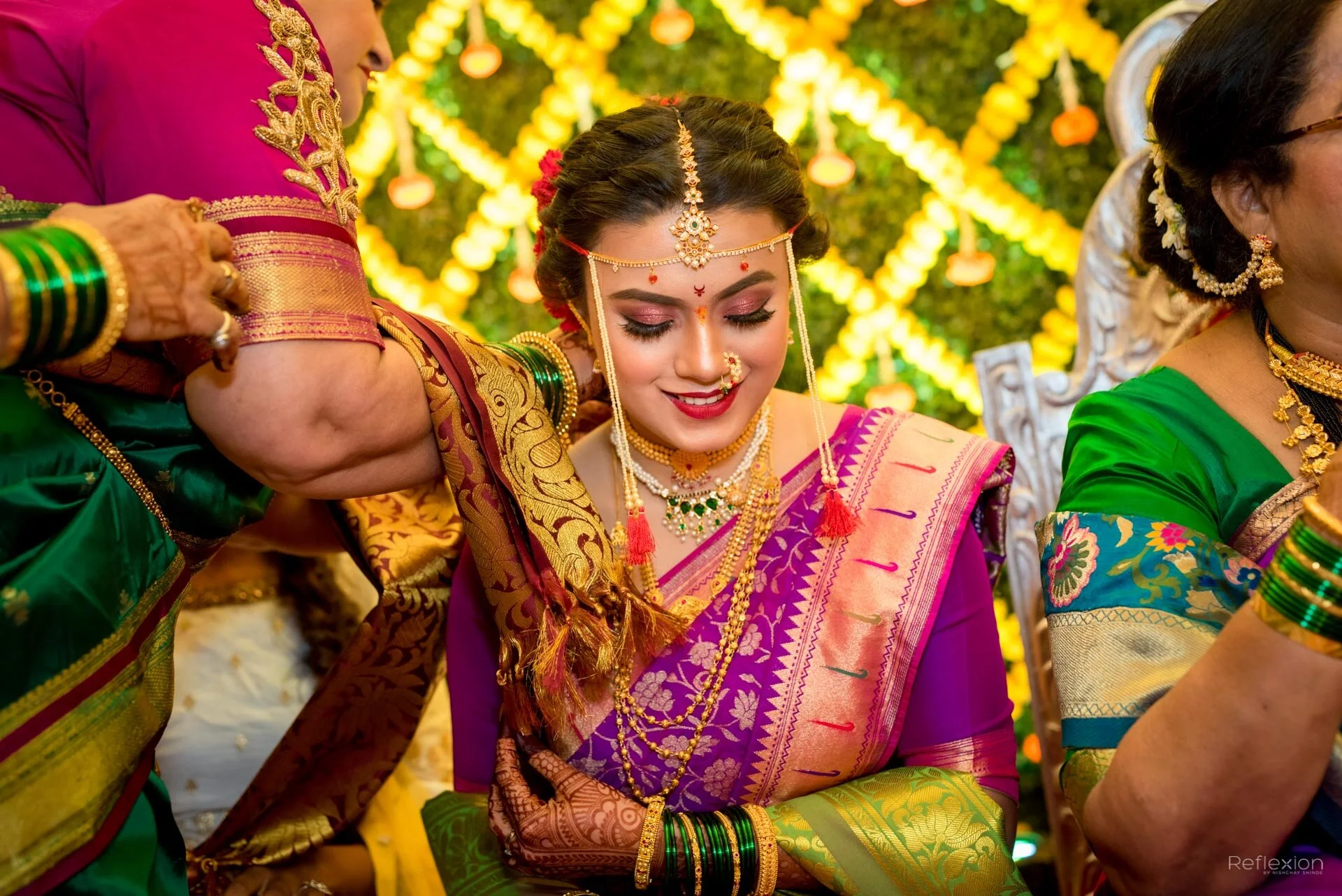 A woman in traditional Indian attire smiling during a celebration, surrounded by women dressed in colorful sarees with intricate jewelry and a decorated backdrop.