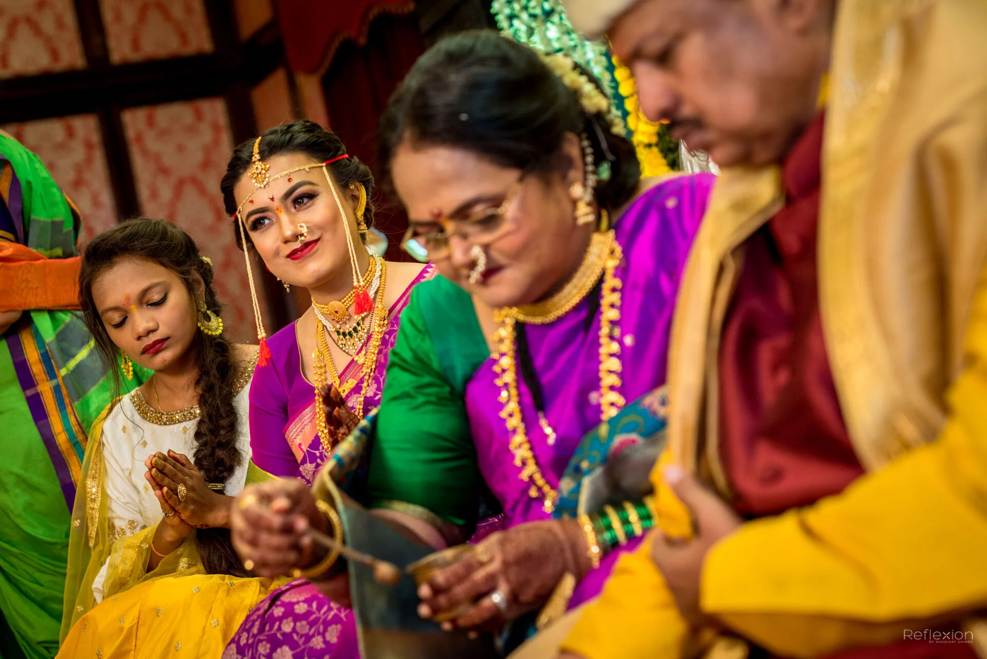People in traditional Indian attire participating in a wedding ceremony, with focus on the bride wearing pink and purple saree and jewelry, and the family members around her.