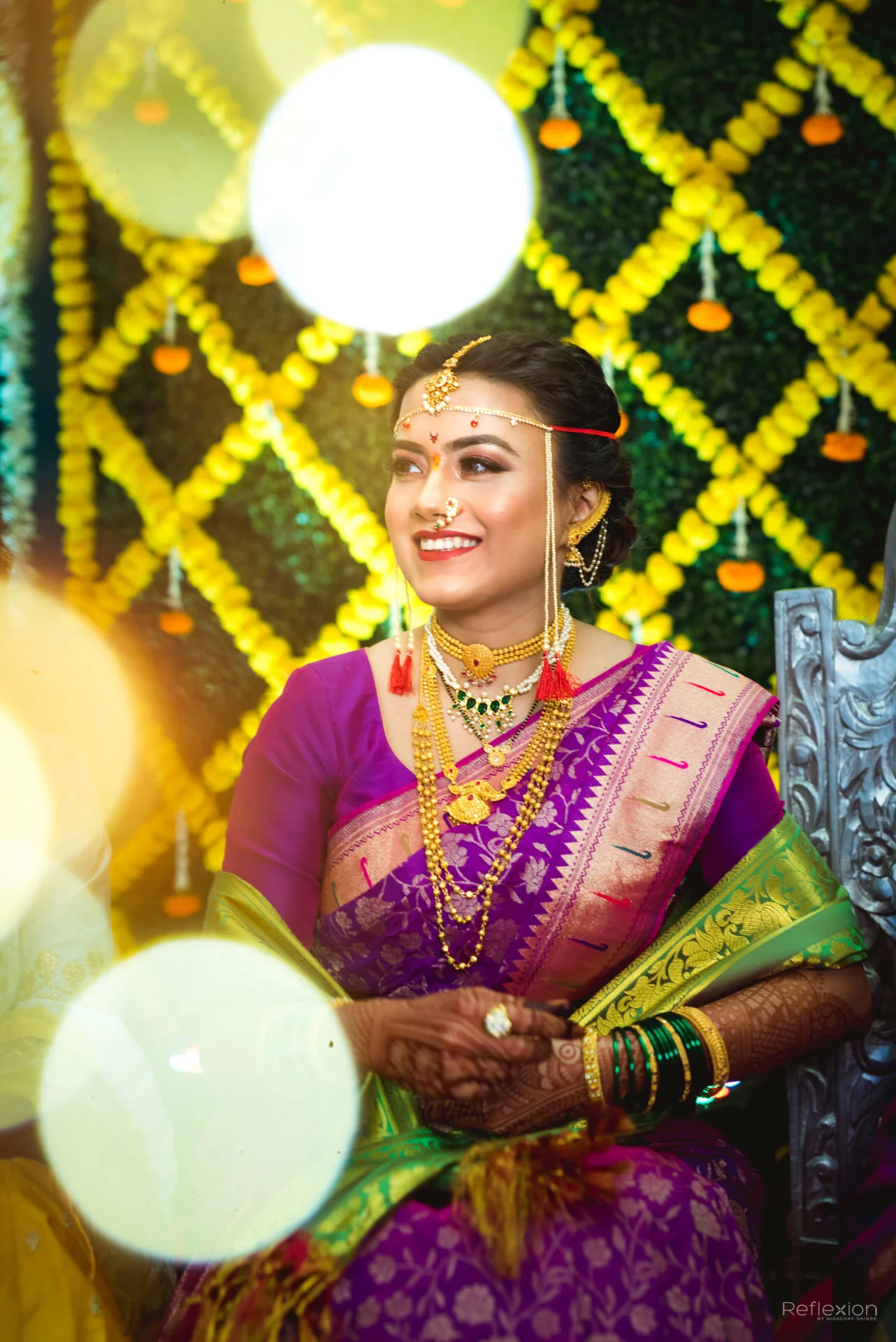 A woman dressed in traditional Indian attire, wearing a purple saree with bright green and gold accents, and adorned with gold jewelry, including necklaces, earrings, a nose ring, and bangles. She is smiling and sitting in front of a vibrant backgrou