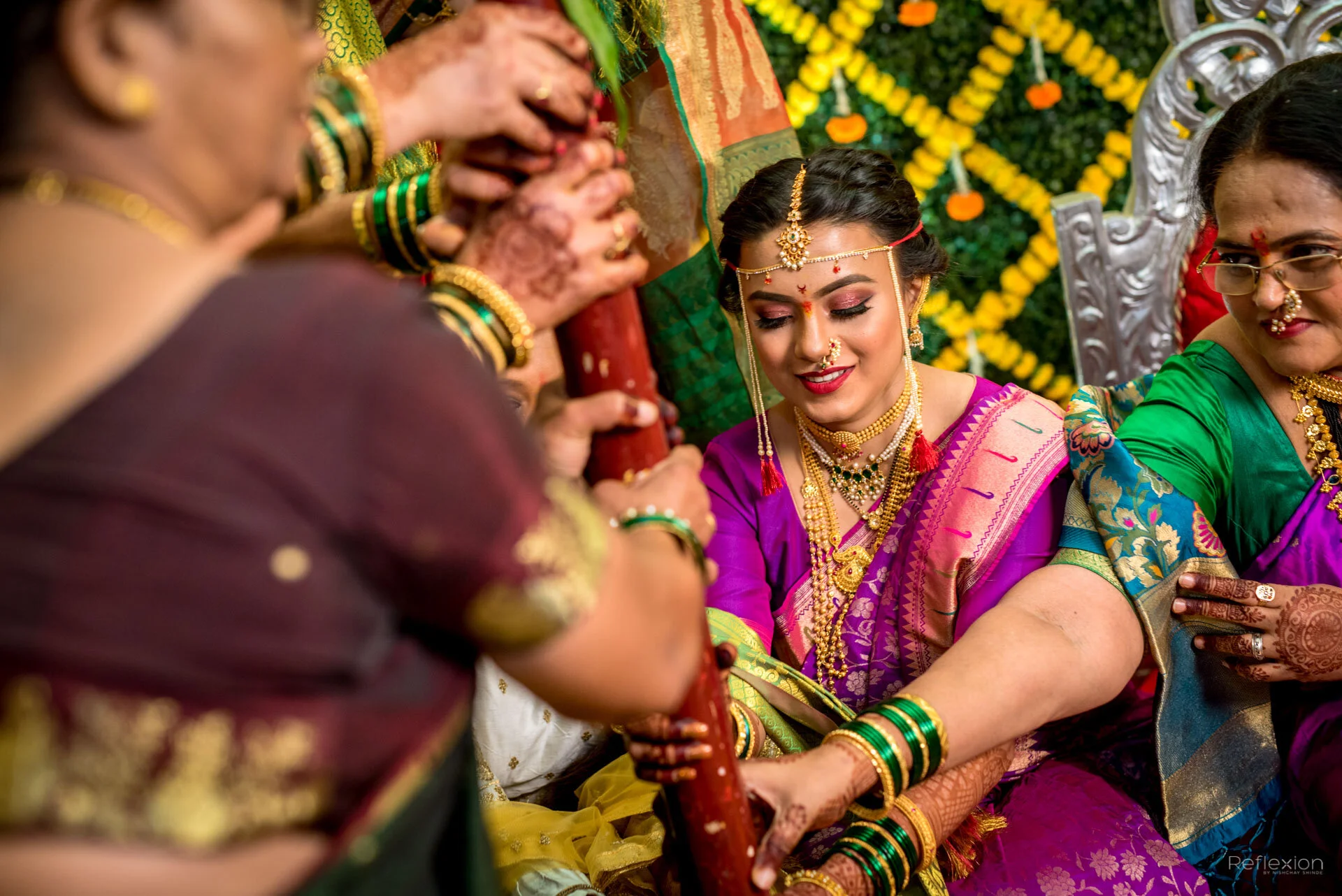 A bride dressed in a vibrant purple and pink saree with gold jewelry, participating in a traditional Indian wedding ceremony, surrounded by women in colorful sarees, holding a wooden stick as part of the ritual, with a decorated floral backdrop.