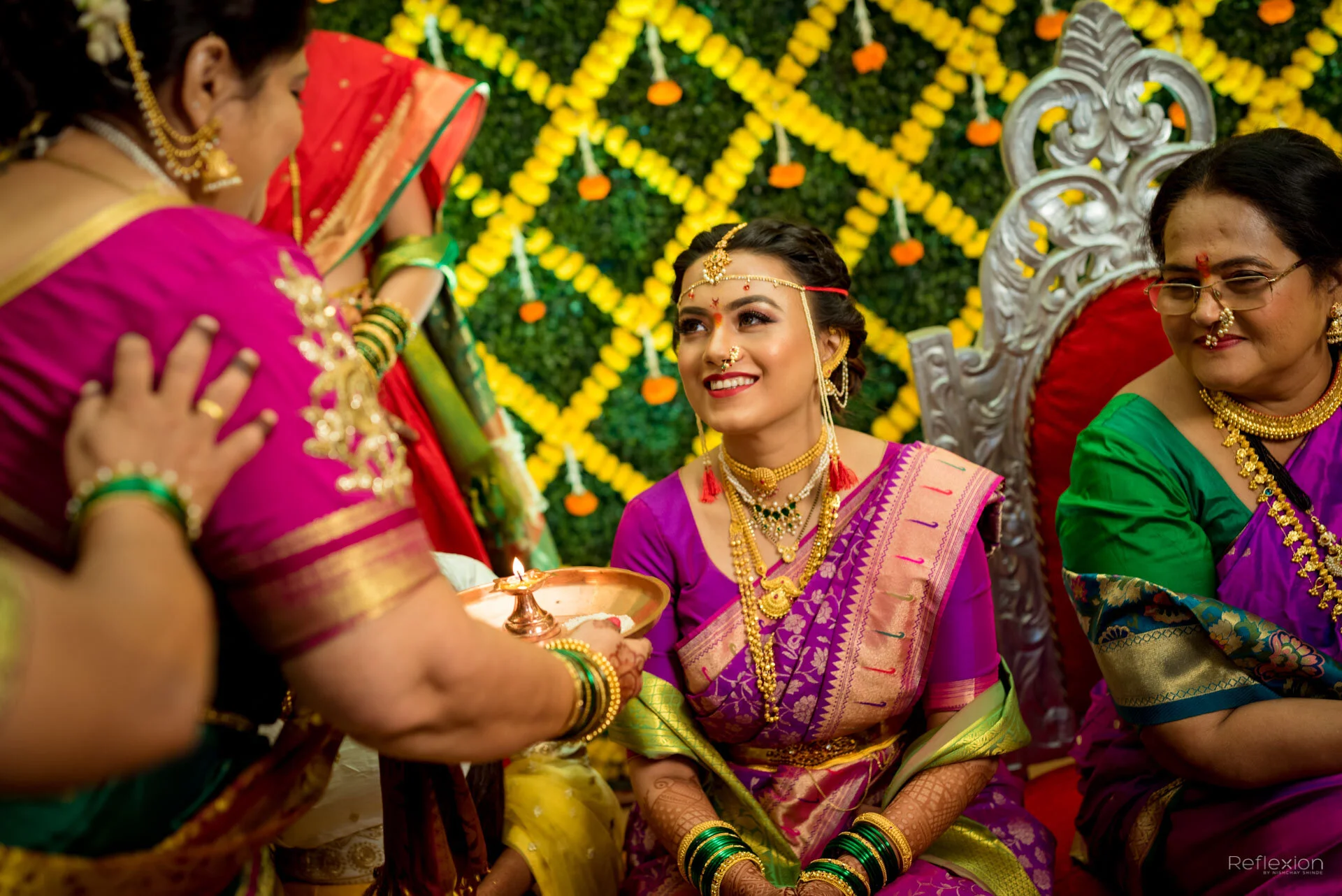 Indian bride wearing traditional purple saree and gold jewelry, sitting on a decorated wedding stage, smiling with women in colorful sarees around her during a wedding ceremony.