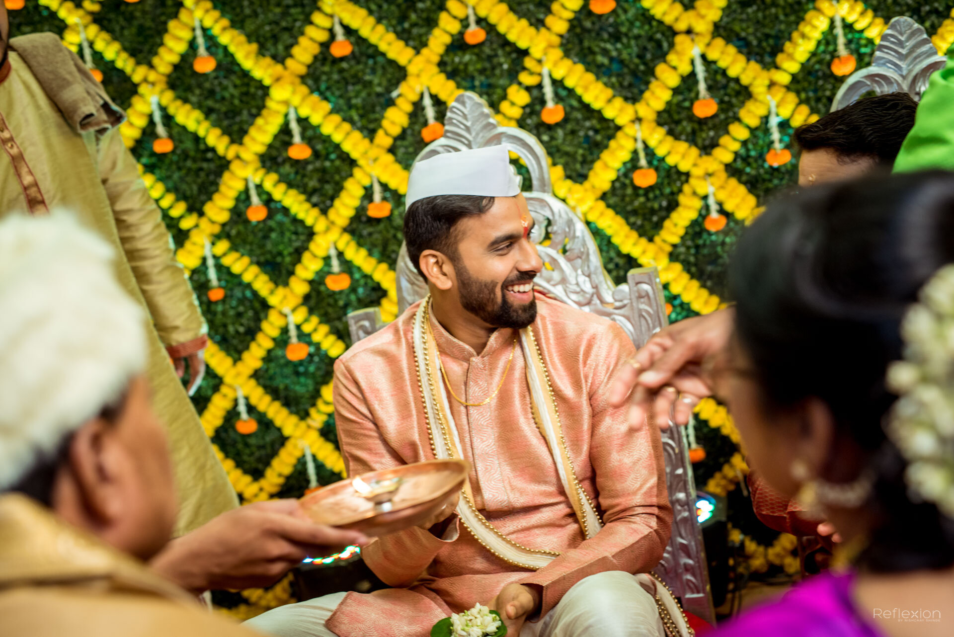 A man dressed in traditional Indian attire, smiling, participating in a wedding ceremony. He is sitting on a decorated silver chair with a yellow and green floral backdrop.