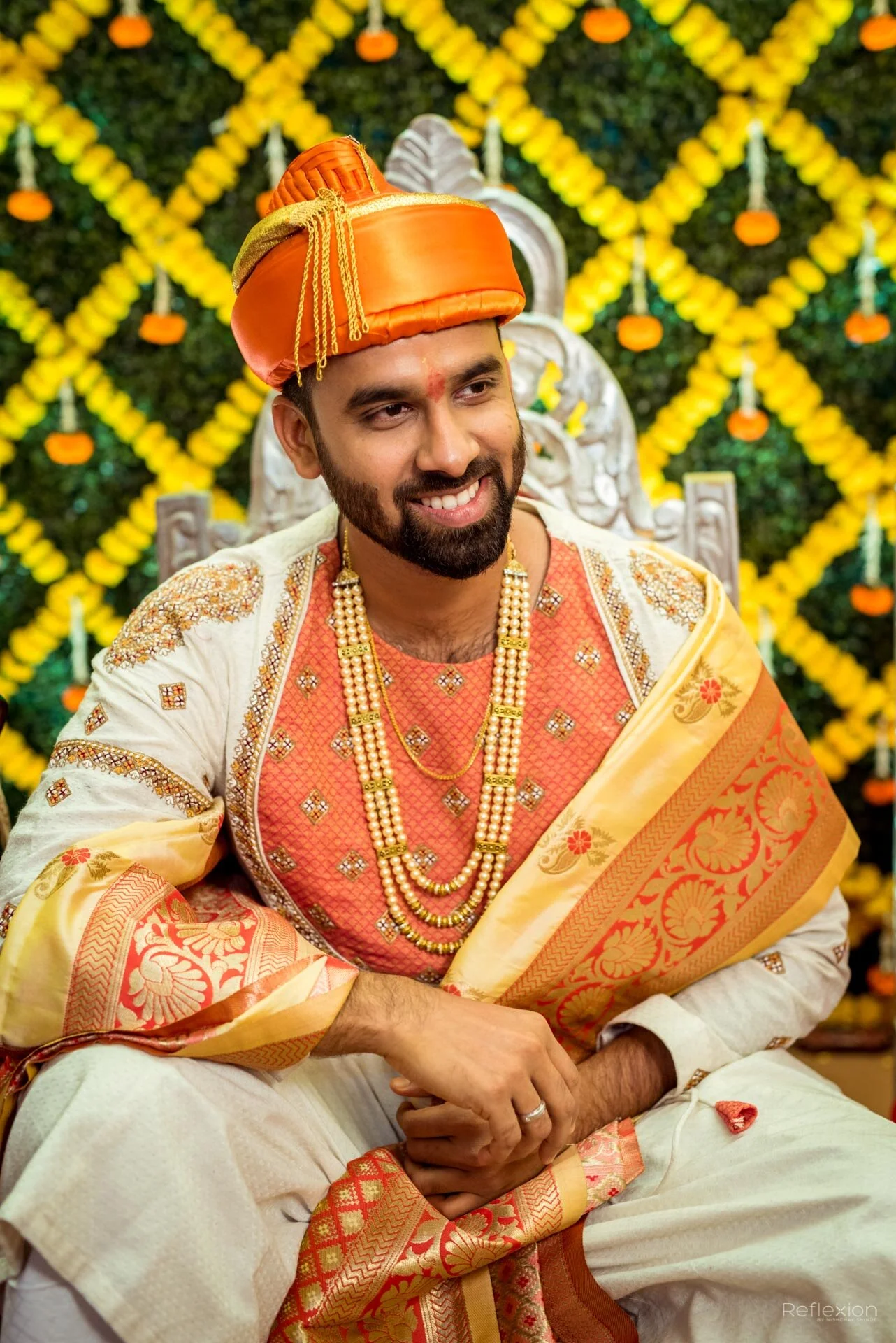 Man dressed in traditional Indian wedding attire, wearing a decorated turban and multiple pearl necklaces, sitting on an ornate chair with decorated floral background.