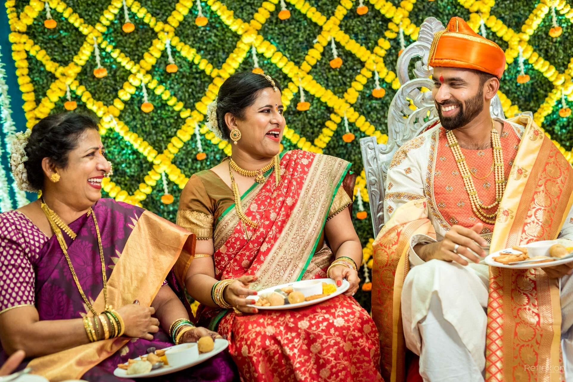 Indian wedding celebration with three women and one man dressed in traditional colorful attire, seated and smiling against a yellow marigold floral backdrop.