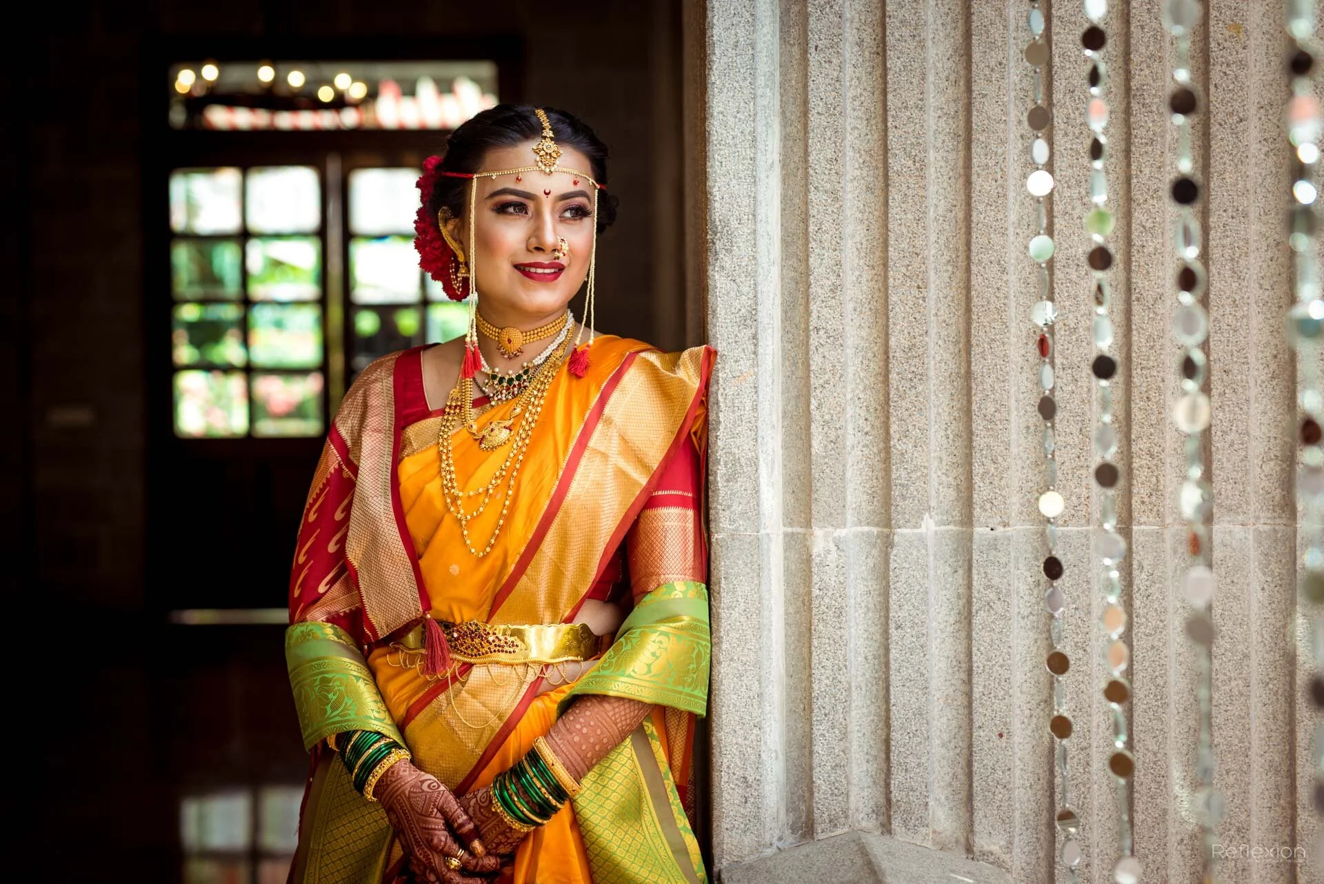 A woman dressed in traditional Indian sari and jewelry, standing near a stone pillar with beaded decorations, indoors with a window in the background.