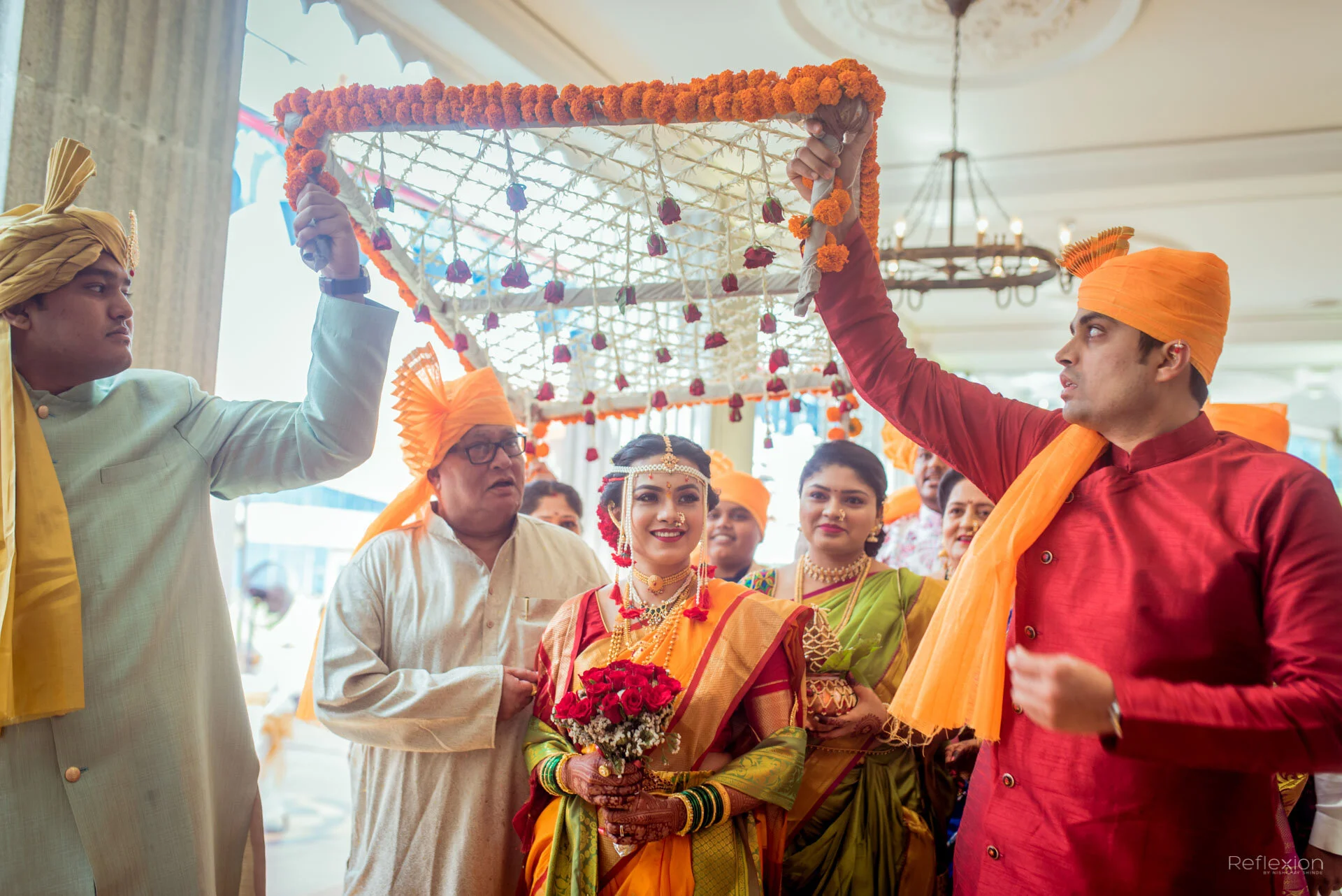 Indian wedding tradition with a bride in a yellow and red saree, holding red roses, surrounded by family and friends in colorful traditional attire, under a decorated canopy.
