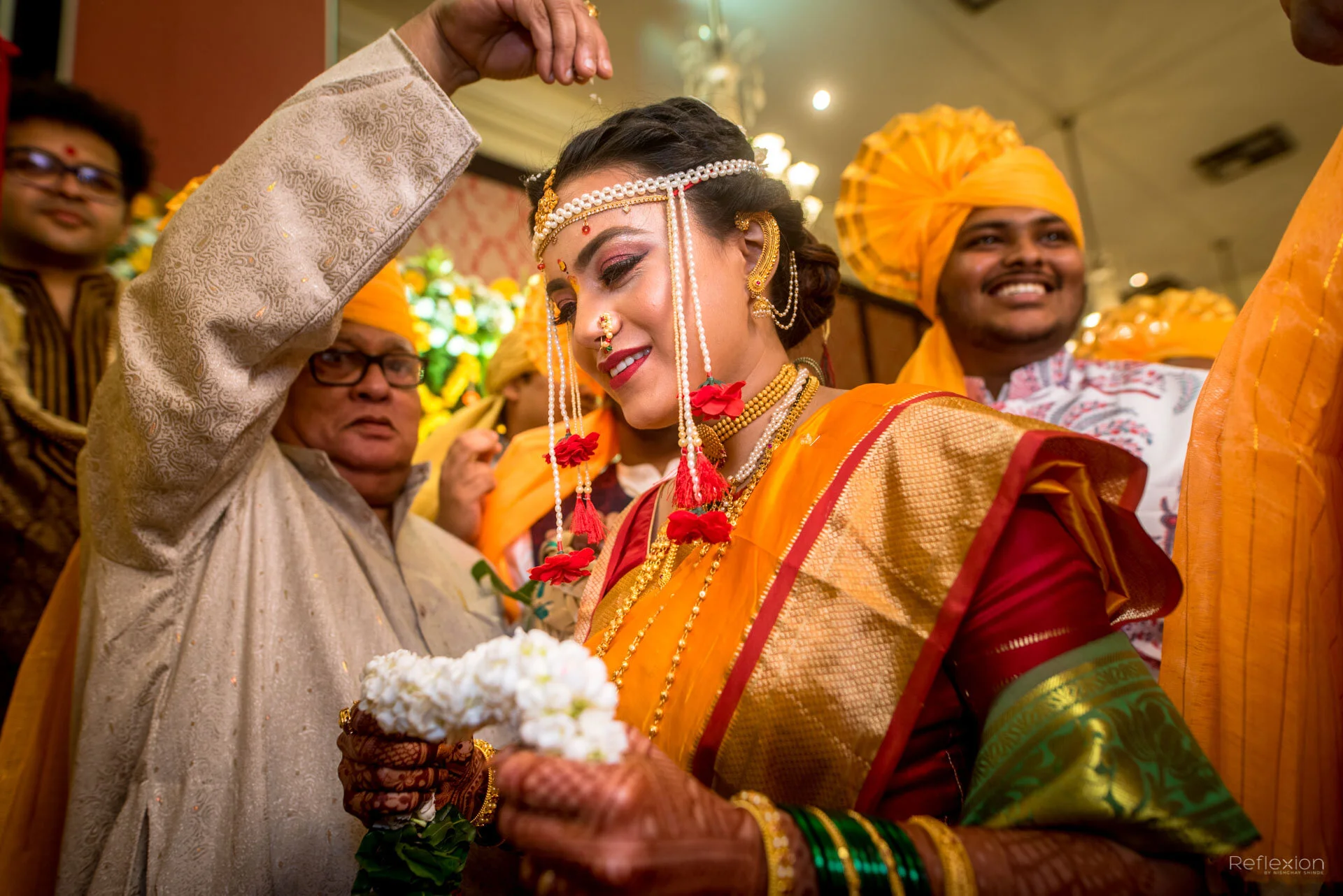 Indian bride dressed in traditional gold and red saree, adorned with jewelry, holding flowers, and surrounded by wedding guests in colorful attire at an Indian wedding ceremony.