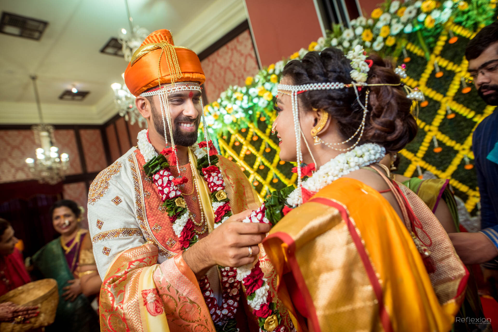 An Indian wedding ceremony with the groom and bride exchanging garlands in traditional attire, smiling at each other, with a decorated background and guests in traditional clothing.