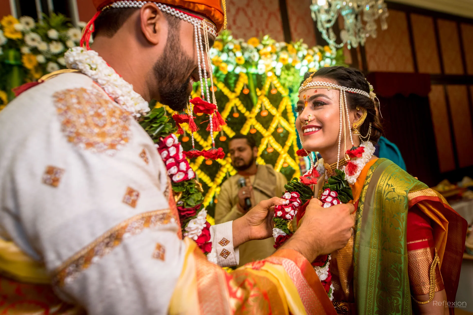 A traditional Indian wedding ceremony where a groom placing a necklace on the smiling bride. Both are dressed in colorful, ornate wedding attire with floral jewelry. The background features bright decorations with yellow and green floral arrangements