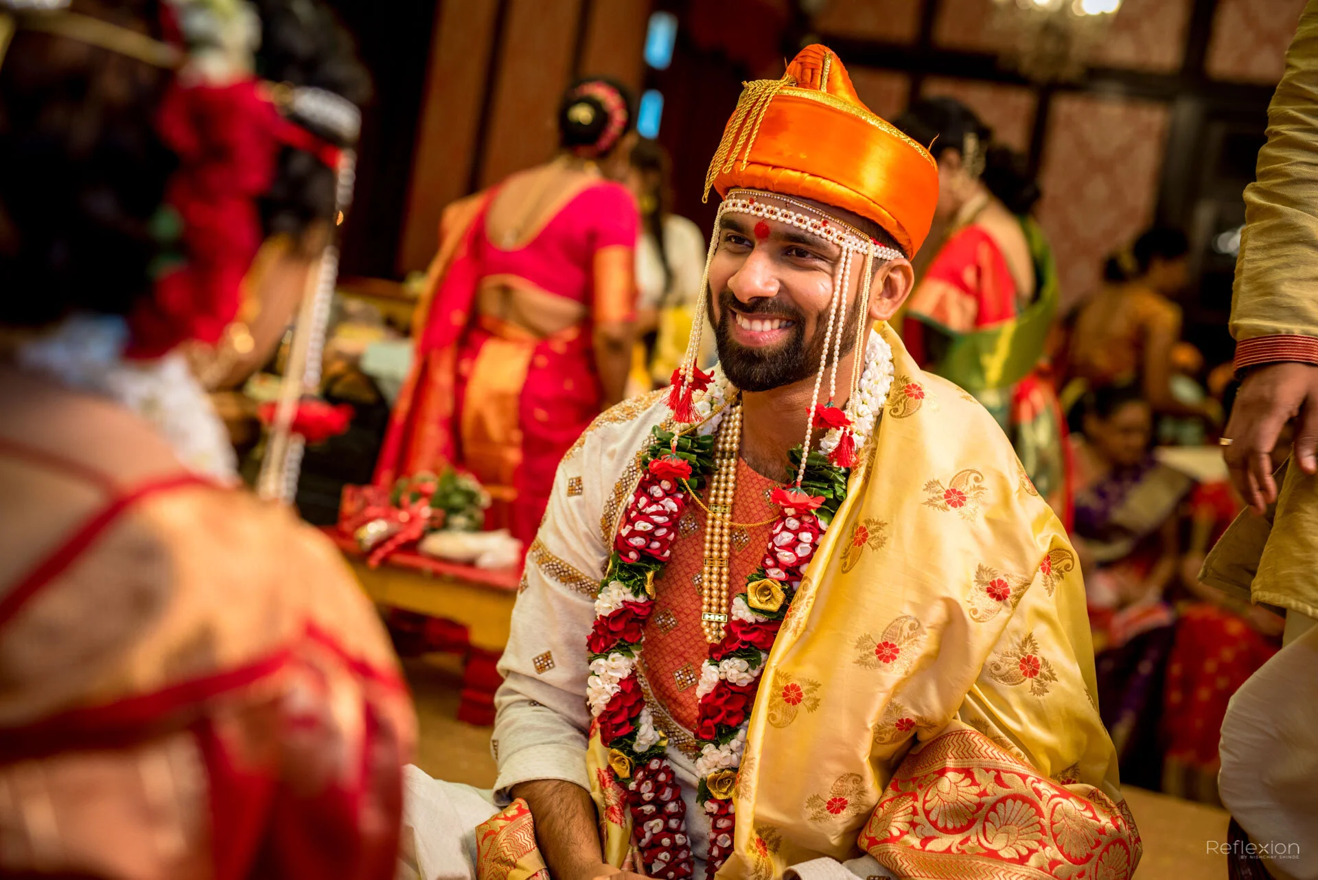 A groom in traditional Indian wedding attire, smiling, wearing a saffron-colored turban, gold embroidery, and adorned with flower garlands, at a colorful wedding ceremony.