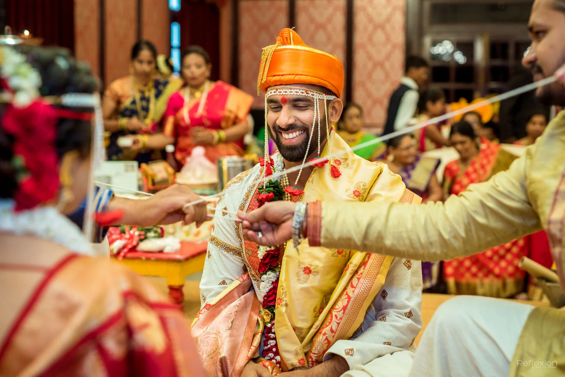 A groom dressed in traditional Indian wedding attire, wearing an orange turban and cream-colored embroidered clothing, is smiling as he participates in a wedding ritual with his bride and relatives, all dressed in colorful traditional Indian clothing