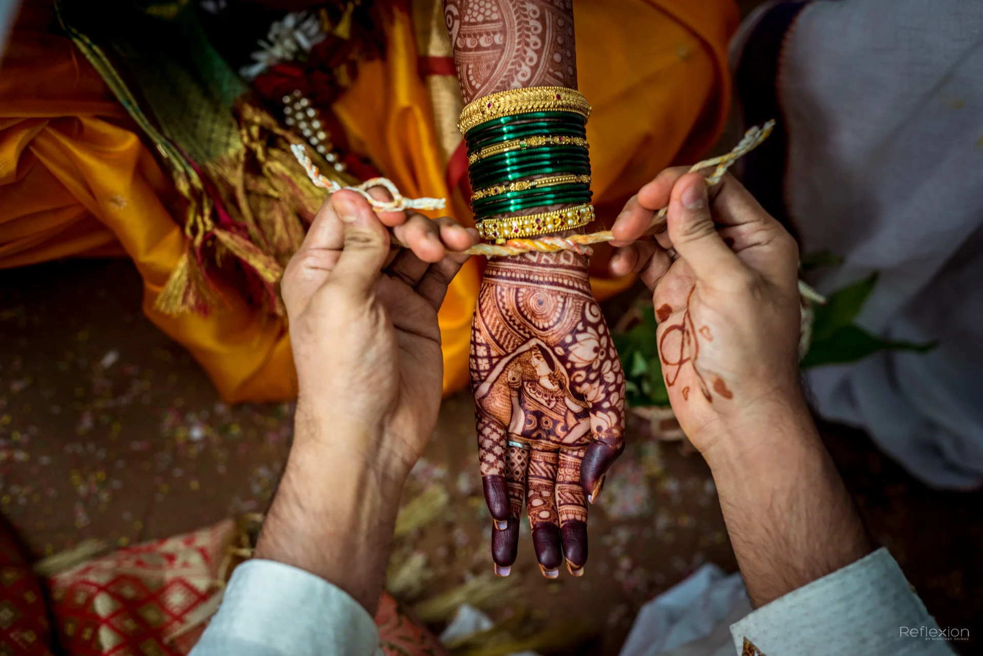 A close-up of a woman's decorated henna hand being tied with a colorful cloth by another person, with woman's arm adorned with green and gold bangles and traditional attire visible.