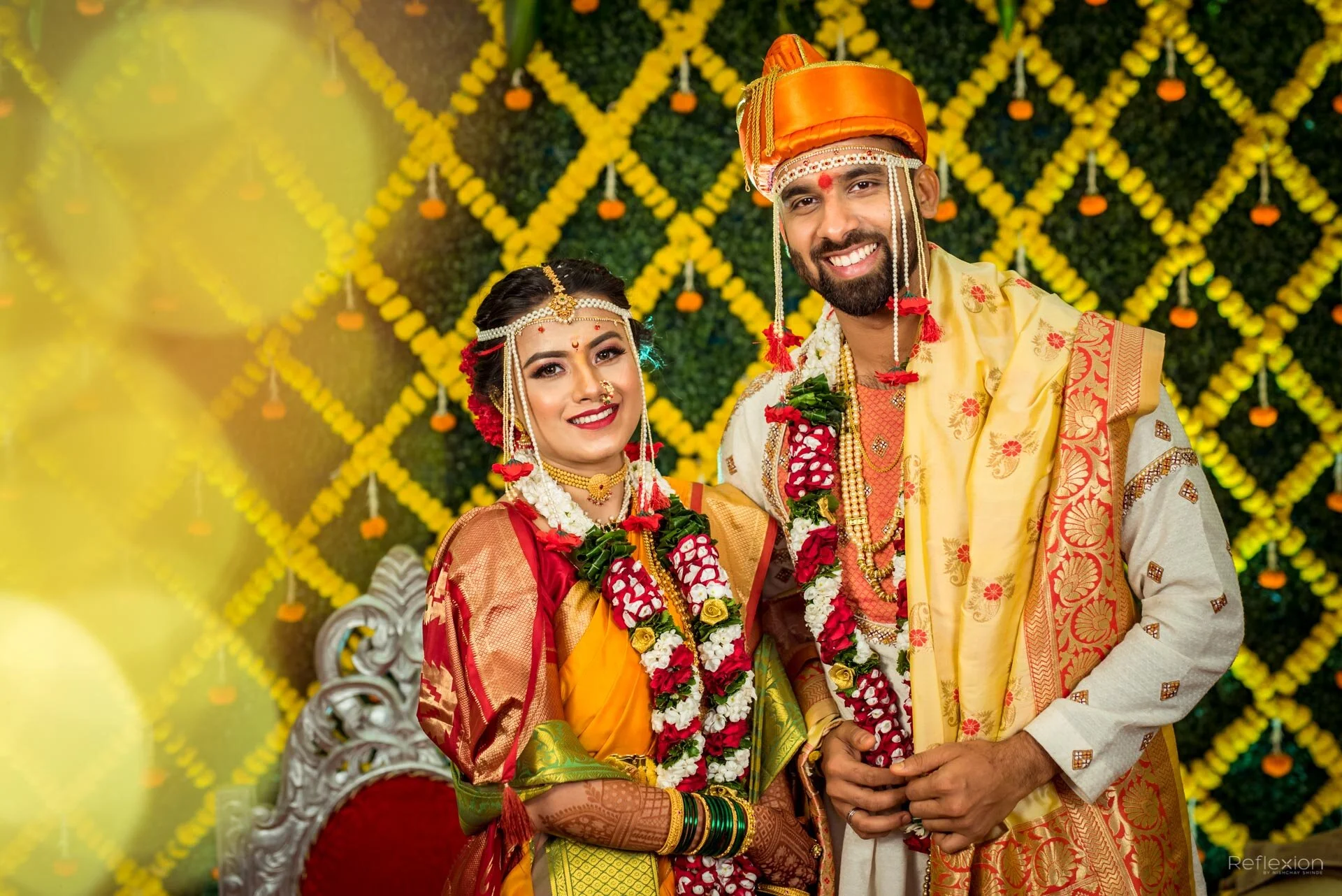 A smiling bride and groom in traditional Indian wedding attire, adorned with jewelry, flowers, and colorful clothing, standing against a lush decorative backdrop of yellow flowers and greenery.