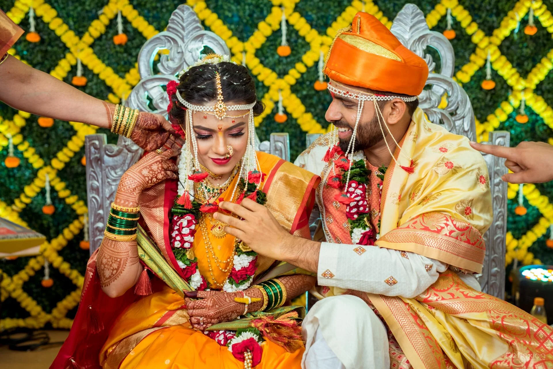 An Indian bride and groom during their wedding ceremony, sitting on a decorated silver throne. The bride is in a red and gold saree with intricate henna on her hands, and the groom is in a cream and gold sherwani with an orange turban. They are excha