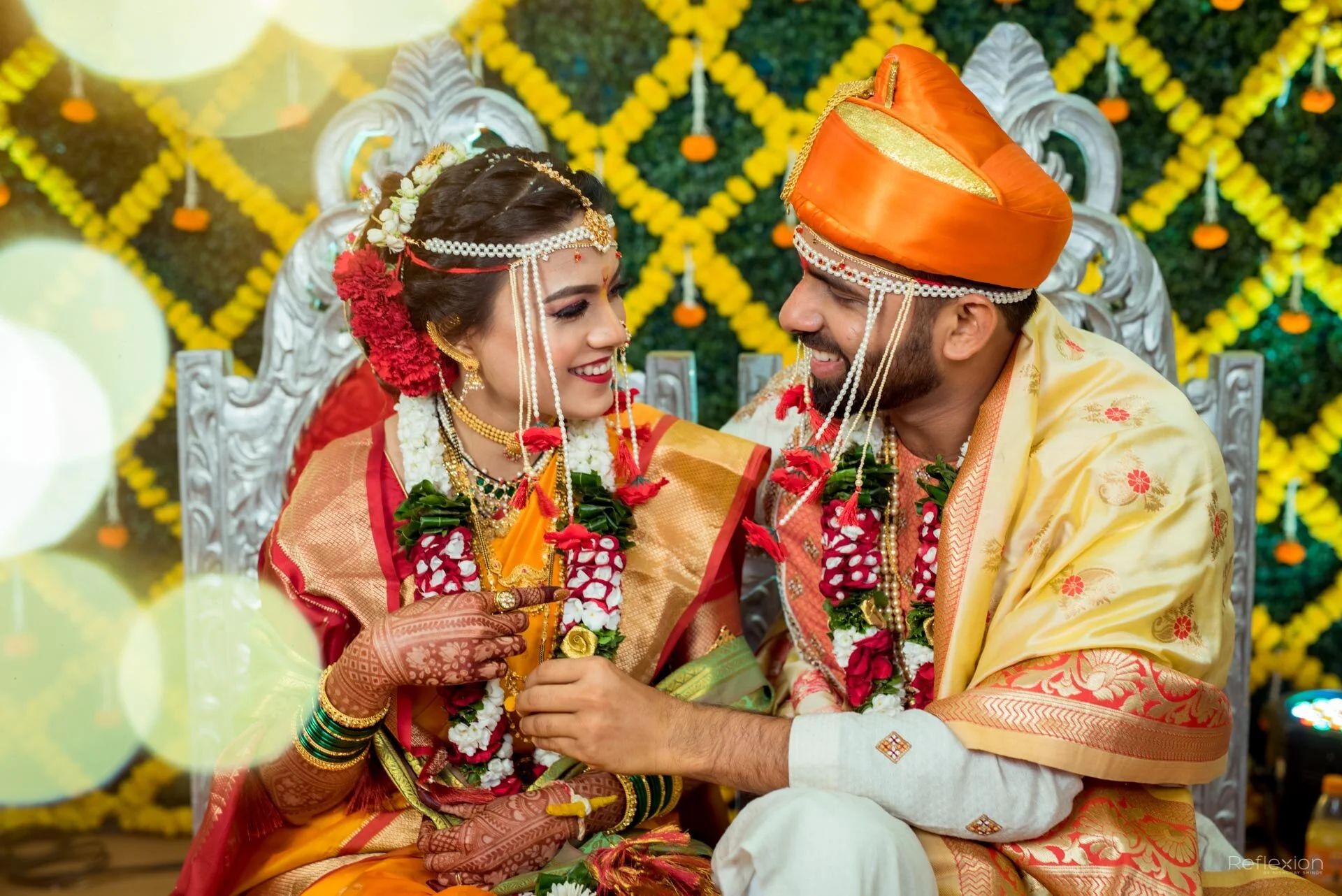 A newlywed Indian couple in traditional wedding attire sits facing each other on a decorated stage, smiling and holding hands, with a backdrop of yellow flowers and green foliage.