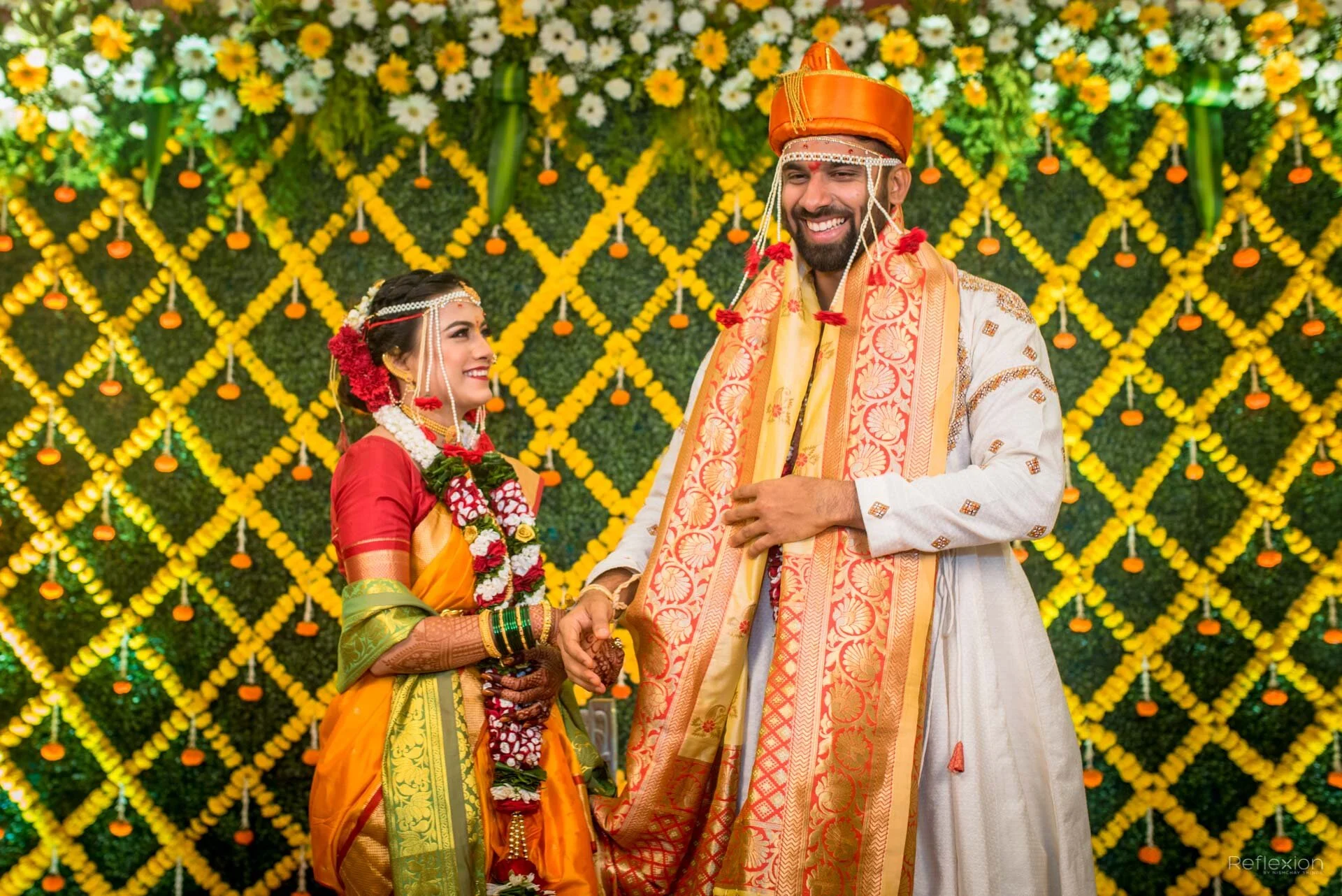 An Indian bride and groom dressed in traditional attire during a wedding ceremony, holding hands and smiling with a decorative yellow and white floral backdrop.