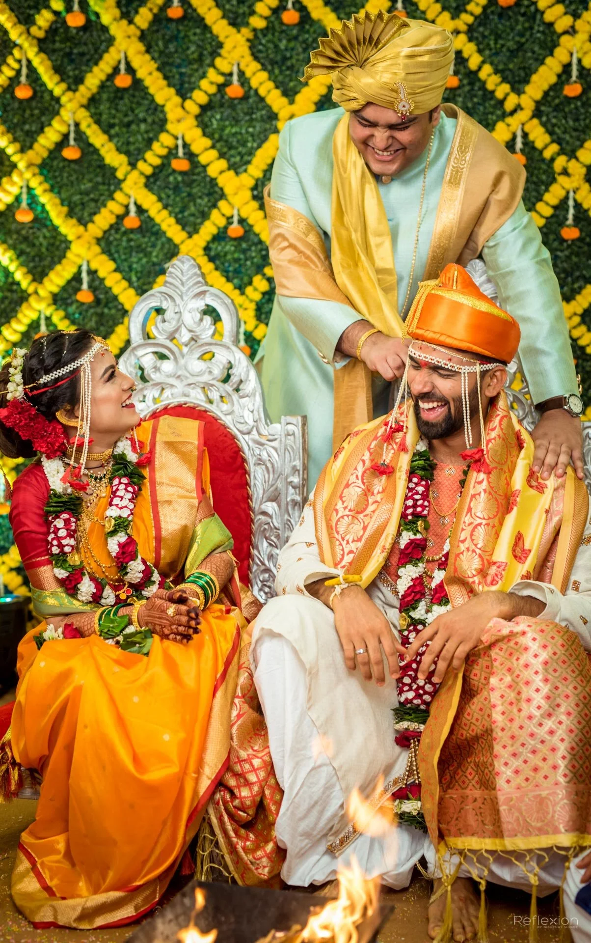 A traditional Indian wedding ceremony with the bride and groom sitting on a decorated stage, exchanging garlands. The bride is dressed in a red and gold saree with jewelry, and the groom in a cream and gold outfit with an orange turban. A man standin