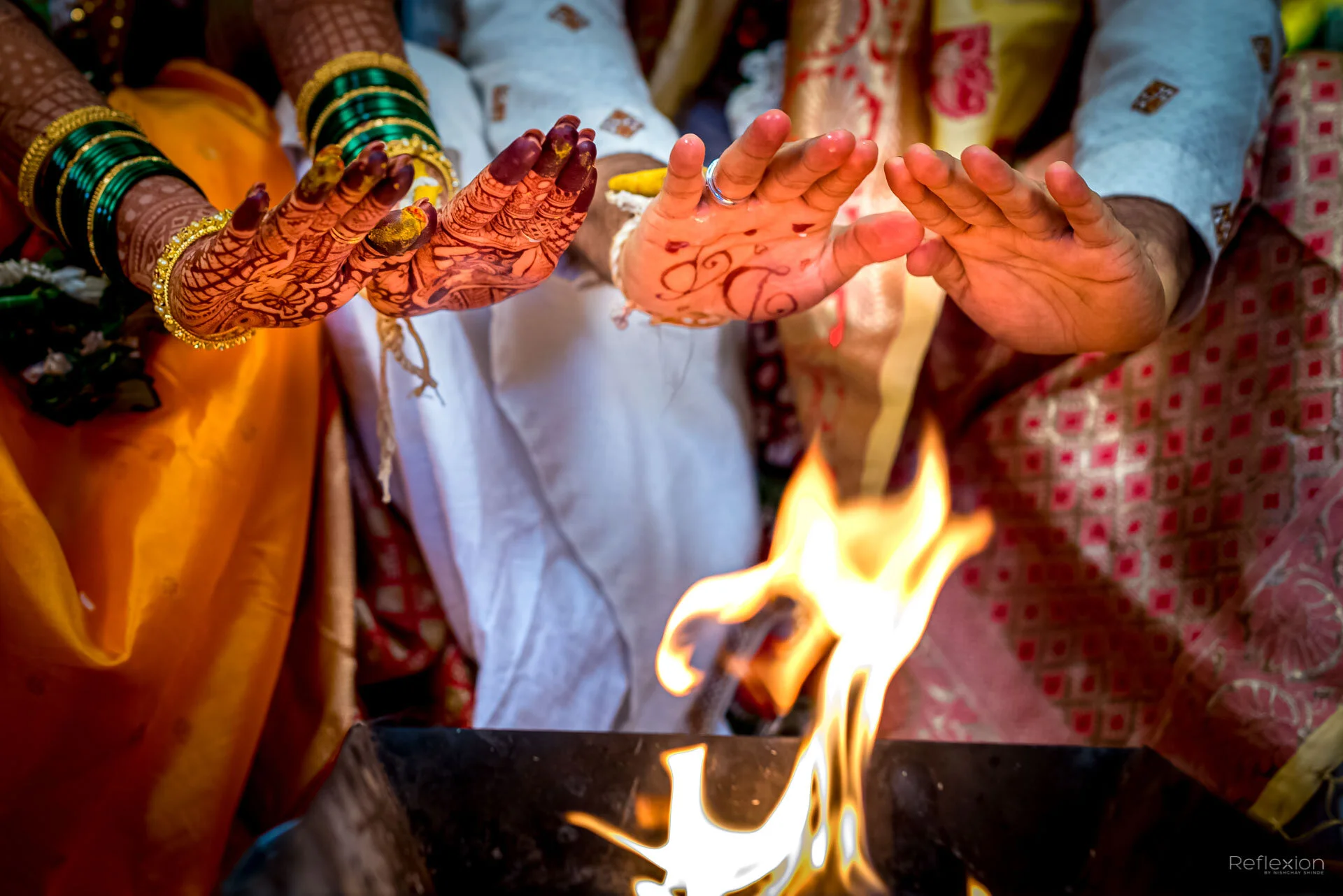 Close-up of hands in traditional Indian wedding ceremony, with intricate henna designs and colorful jewelry, near a sacred fire.